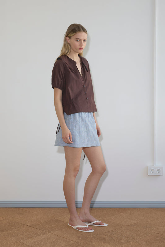 A young woman with long blonde hair stands indoors against a plain wall, wearing the Double Collar Shirt - Umber by Deiji Studios, a light blue skirt, and white sandals. She looks slightly to the side with a neutral expression.