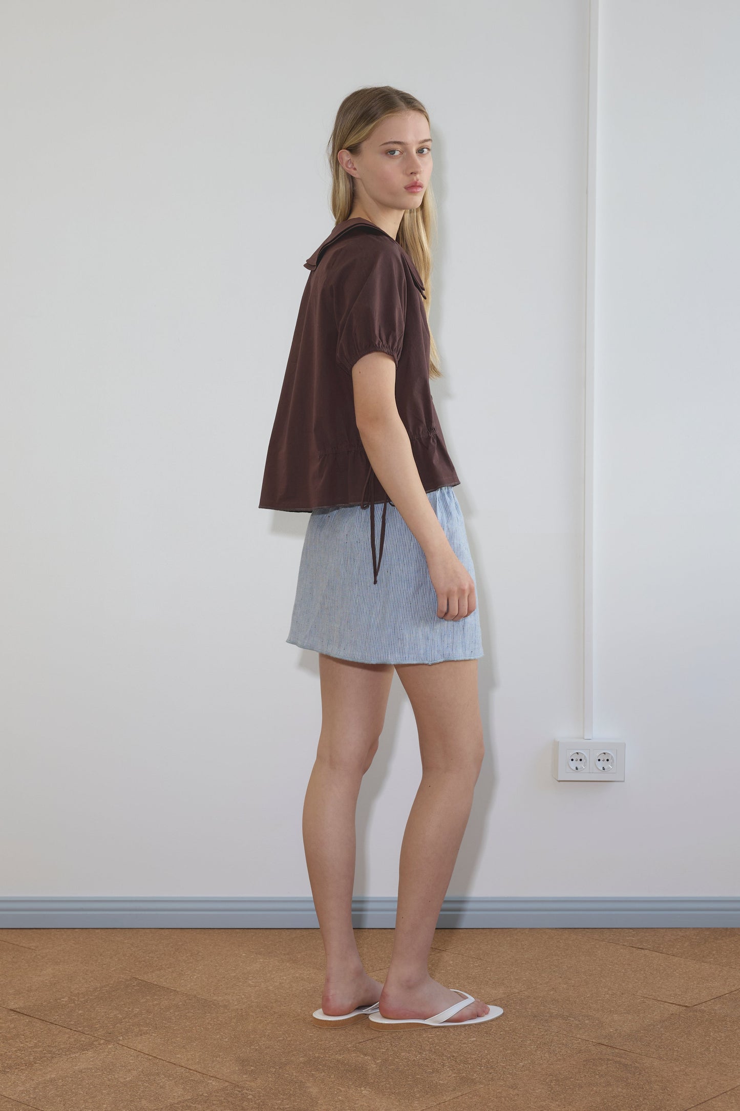 A woman with long blonde hair stands sideways in a room, wearing the Umber Double Collar Shirt by Deiji Studios, a light blue skirt, and white slide sandals. The background includes a white wall, power outlet, and brown floor.