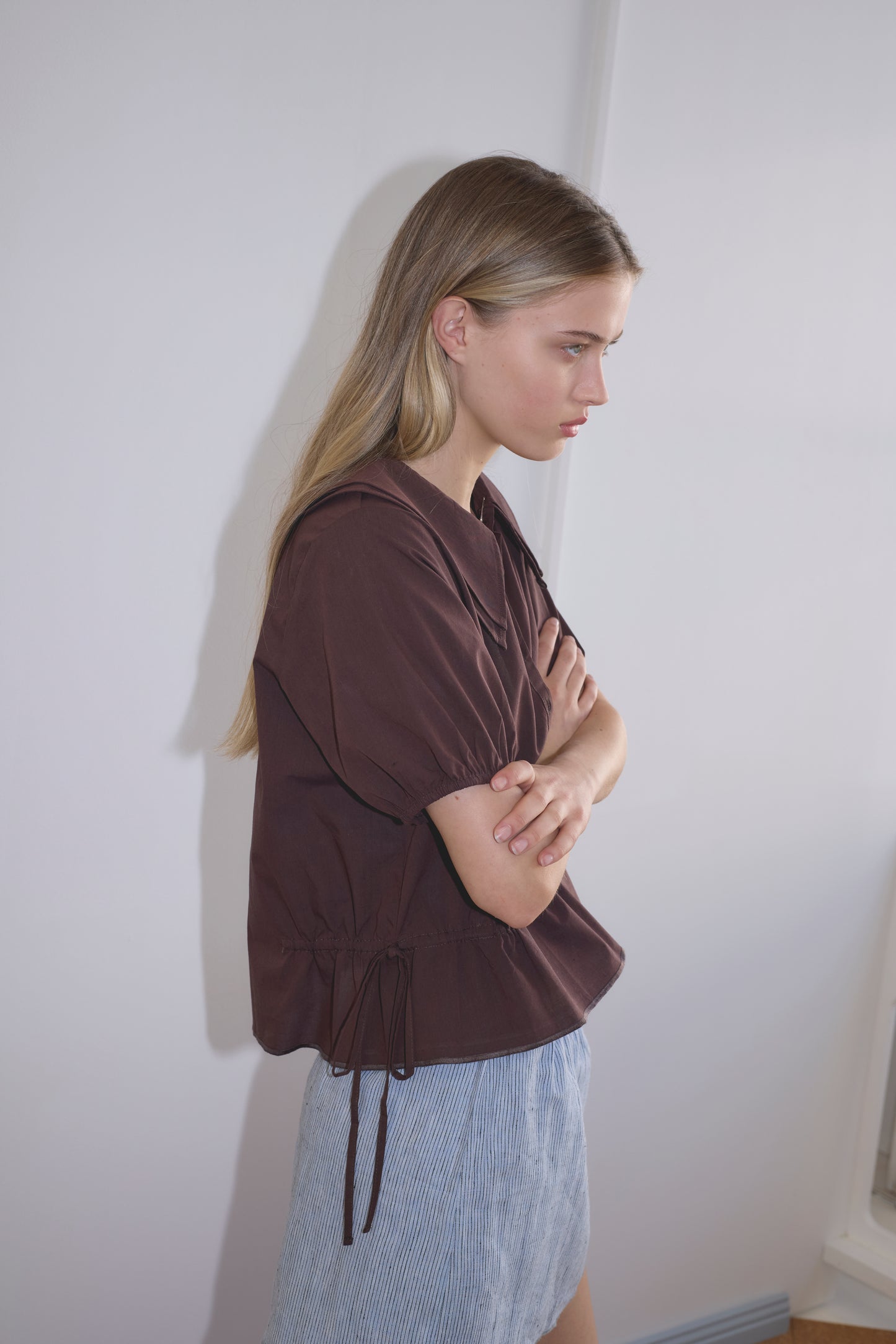 A young woman with long blonde hair stands sideways, arms crossed, wearing the Double Collar Shirt - Umber by Deiji Studios and a light blue skirt. She appears thoughtful, looking slightly downward against a light-colored wall.