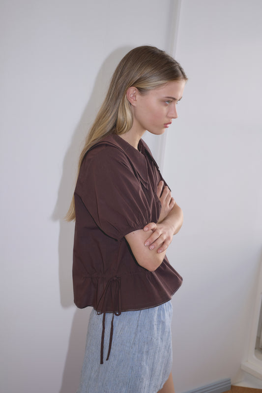 A young woman with long blonde hair stands sideways, arms crossed, wearing the Double Collar Shirt - Umber by Deiji Studios and a light blue skirt. She appears thoughtful, looking slightly downward against a light-colored wall.