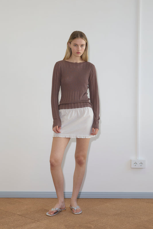 A woman with straight light brown hair and a neutral expression stands indoors against a white wall, wearing the Flounce Mini - Gull Stripe skirt by Deiji Studios and a long-sleeved brown top. An electrical outlet is visible on the wall nearby.