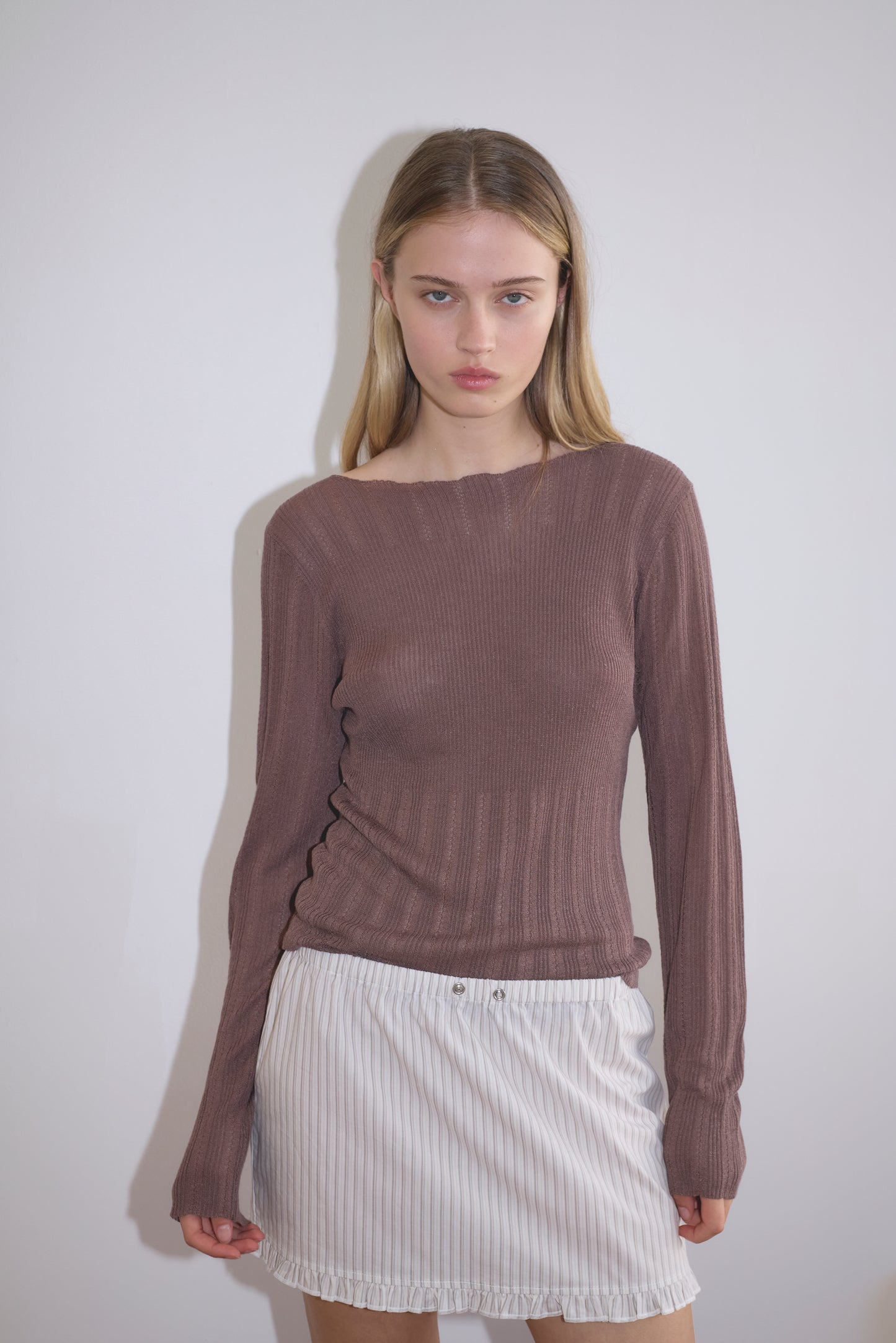 A young woman with straight blonde hair stands against a white wall, wearing the Flounce Mini - Gull Stripe, a white pinstriped skirt, paired with a long-sleeved ribbed brown top. She looks directly at the camera with a neutral expression.