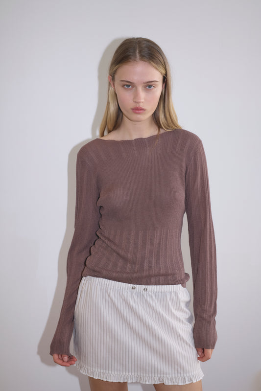 A young woman with straight blonde hair stands against a white wall, wearing the Flounce Mini - Gull Stripe, a white pinstriped skirt, paired with a long-sleeved ribbed brown top. She looks directly at the camera with a neutral expression.