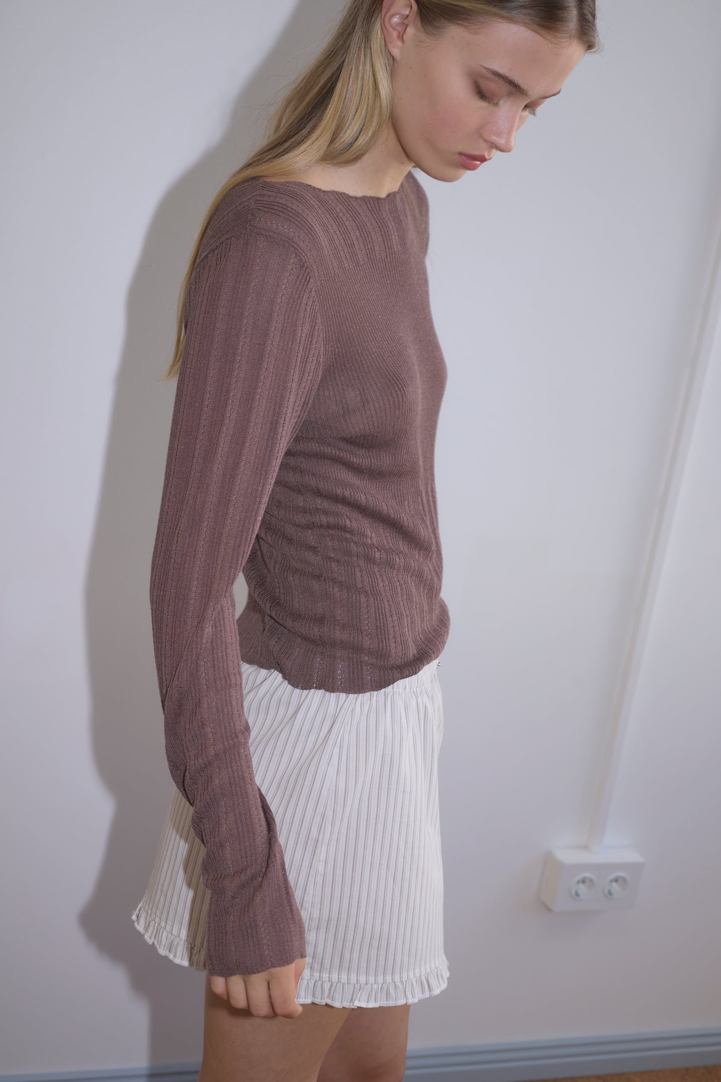 A woman with long blonde hair stands sideways in a ribbed mauve top and the Flounce Mini - Gull Stripe skirt from Deiji Studios, against a plain white wall with an electrical outlet visible in the background.