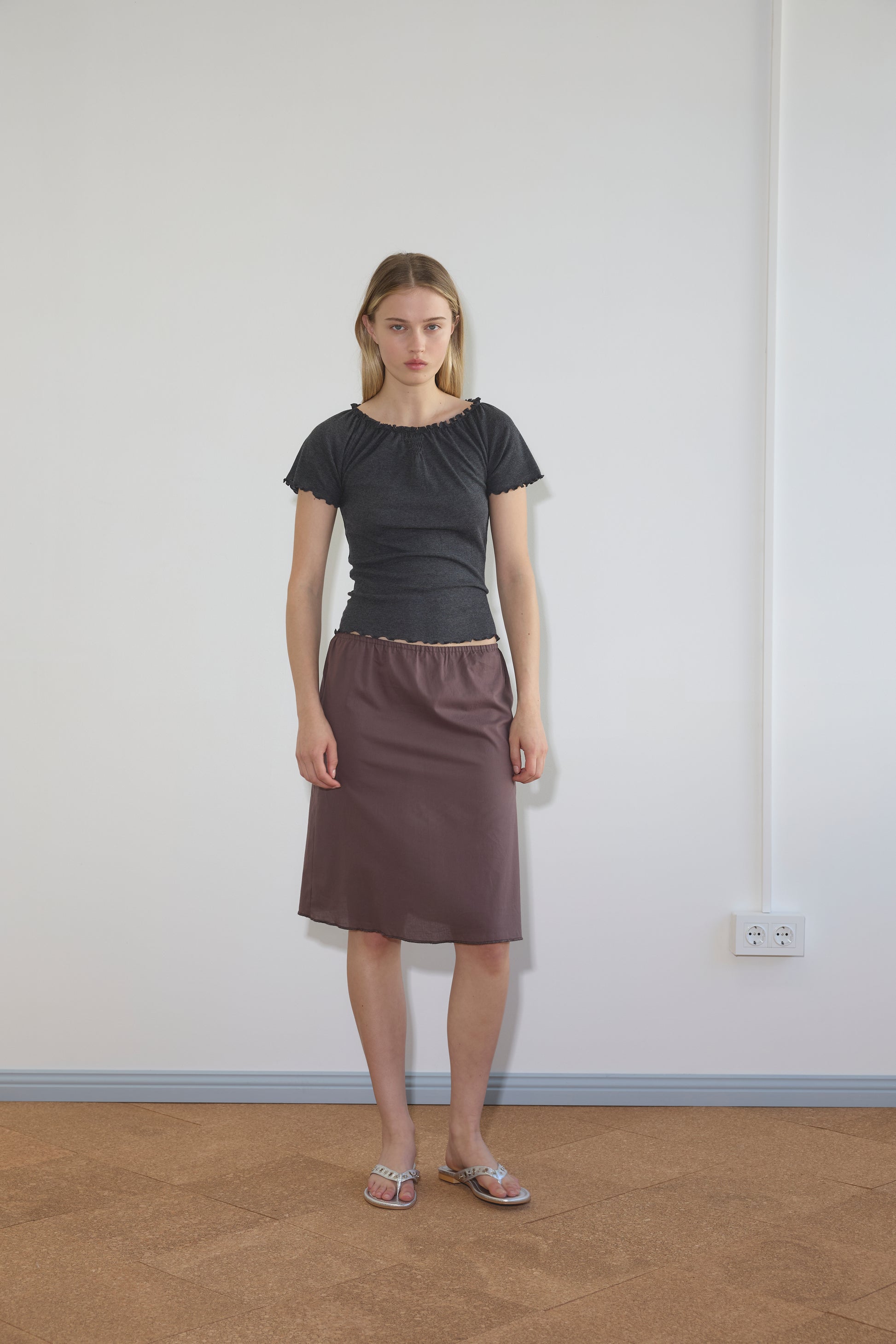 A woman with straight blonde hair and a neutral expression stands indoors against a white wall, wearing a dark short-sleeved top, sandals, and the Simple Skirt - Raisin. An electrical outlet appears on the wall behind her.