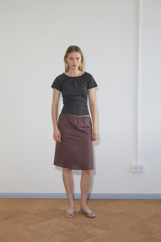 A woman with straight blonde hair and a neutral expression stands indoors against a white wall, wearing a dark short-sleeved top, sandals, and the Simple Skirt - Raisin. An electrical outlet appears on the wall behind her.