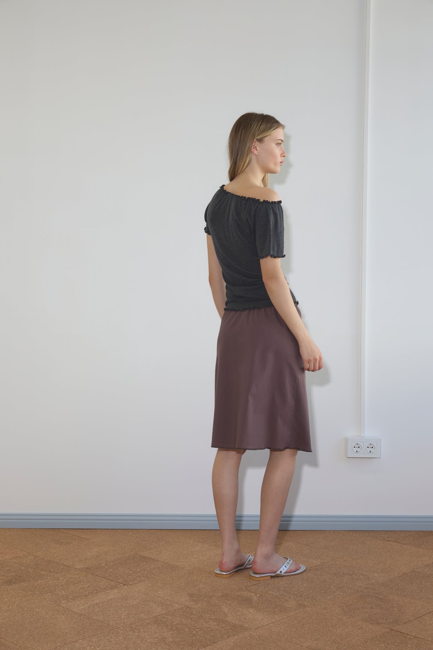 A woman stands indoors facing a white wall, wearing a black top, the Simple Skirt - Raisin, and pink sandals. The room has a light floor, an electrical outlet, and visible wall wiring.
