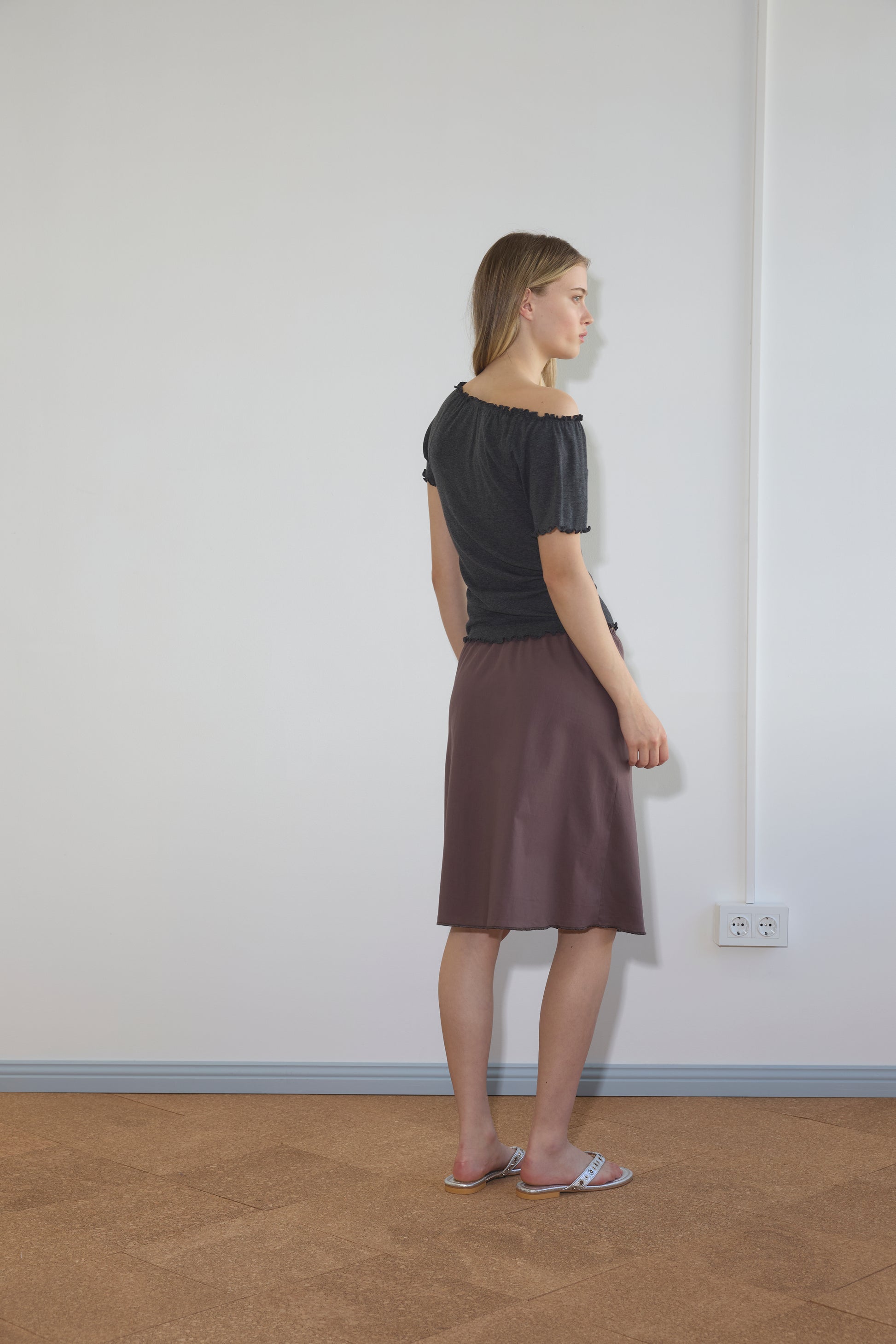 A woman stands indoors facing a white wall, wearing a black top, the Simple Skirt - Raisin, and pink sandals. The room has a light floor, an electrical outlet, and visible wall wiring.