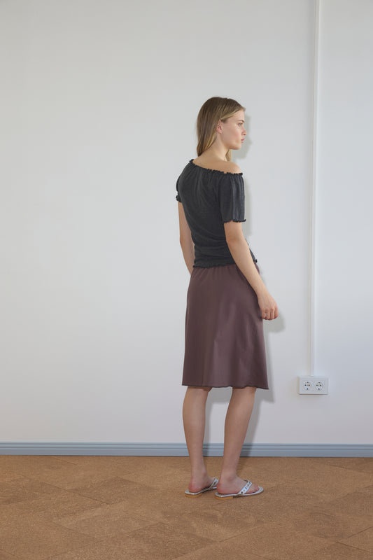 A woman stands indoors facing a white wall, wearing a black top, the Simple Skirt - Raisin, and pink sandals. The room has a light floor, an electrical outlet, and visible wall wiring.