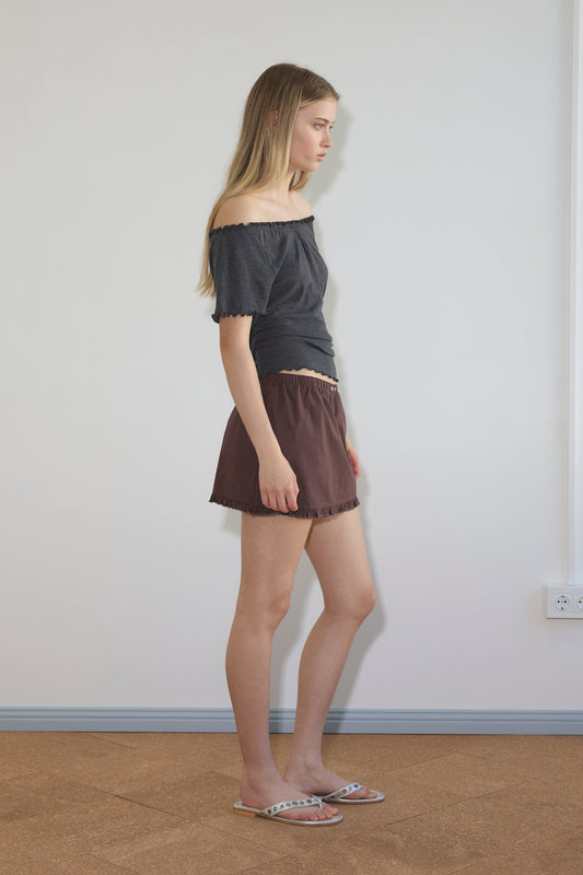 A woman stands in profile, wearing a dark gray off-the-shoulder top, Deiji Studios’ Flounce Mini - Umber organic cotton skirt, and sandals with white straps and multicolored dots. She has long, straight hair and a neutral expression.