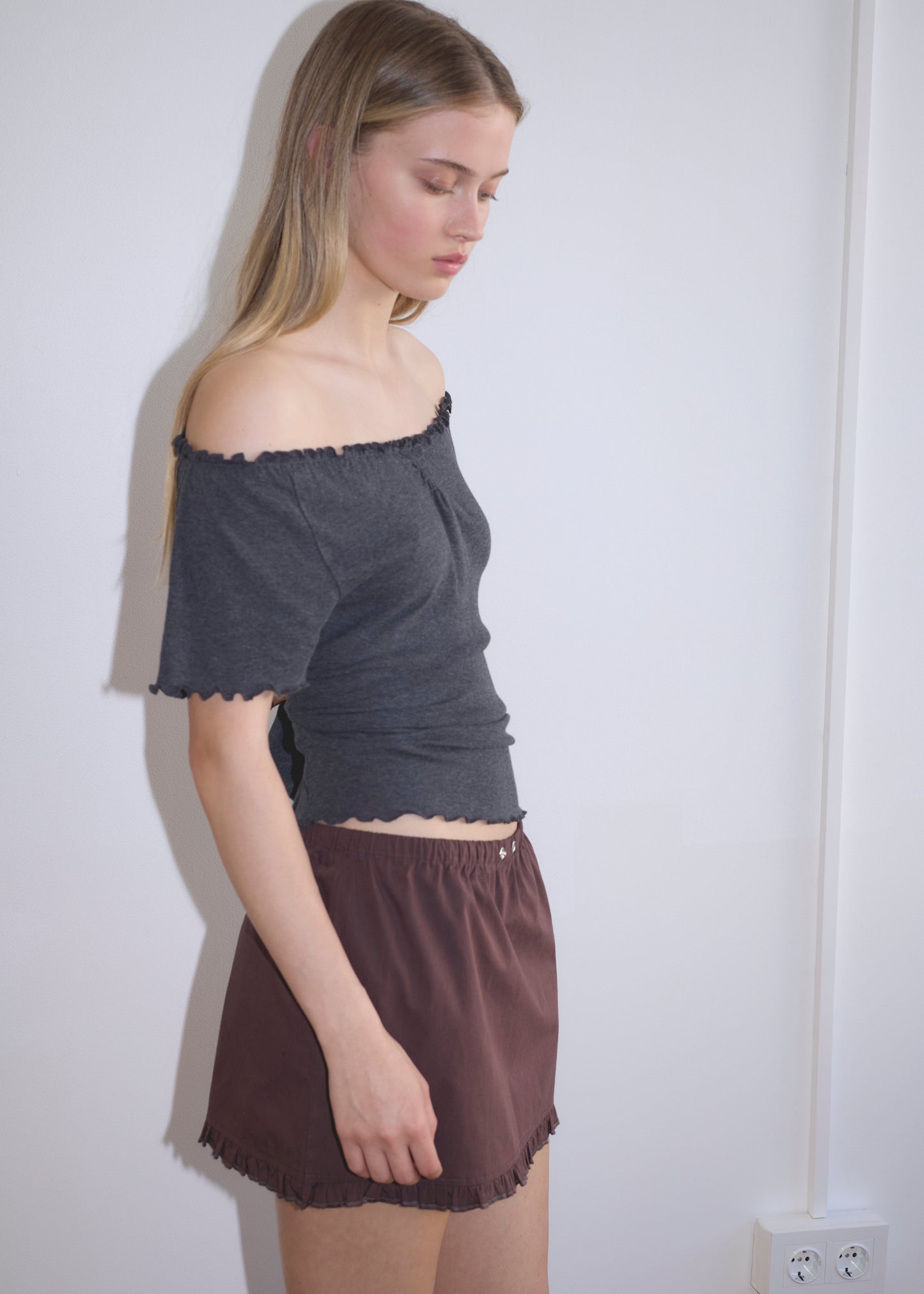 A young woman with long blonde hair stands sideways, looking down. She wears an off-the-shoulder dark gray top and the Flounce Mini - Umber skirt, set against a plain white wall with a visible power outlet near the floor.
