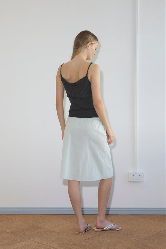 A woman indoors faces a white wall, wearing a black sleeveless top, sandals, and the Simple Skirt - Mint by Deiji Studios. A power outlet is visible on the wall nearby.