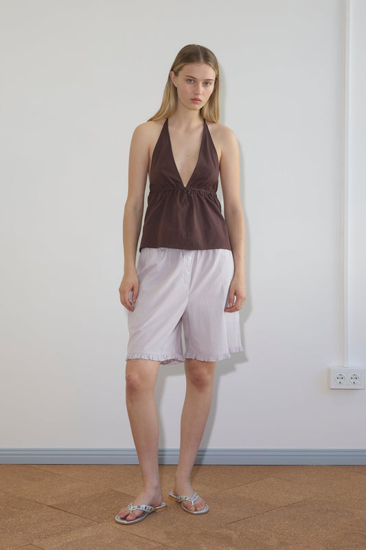 A woman stands indoors against a plain wall wearing a brown halter top, Flounce Short - Lilac Stripe by Deiji Studios, and sandals. She has straight, long blonde hair and a neutral expression.