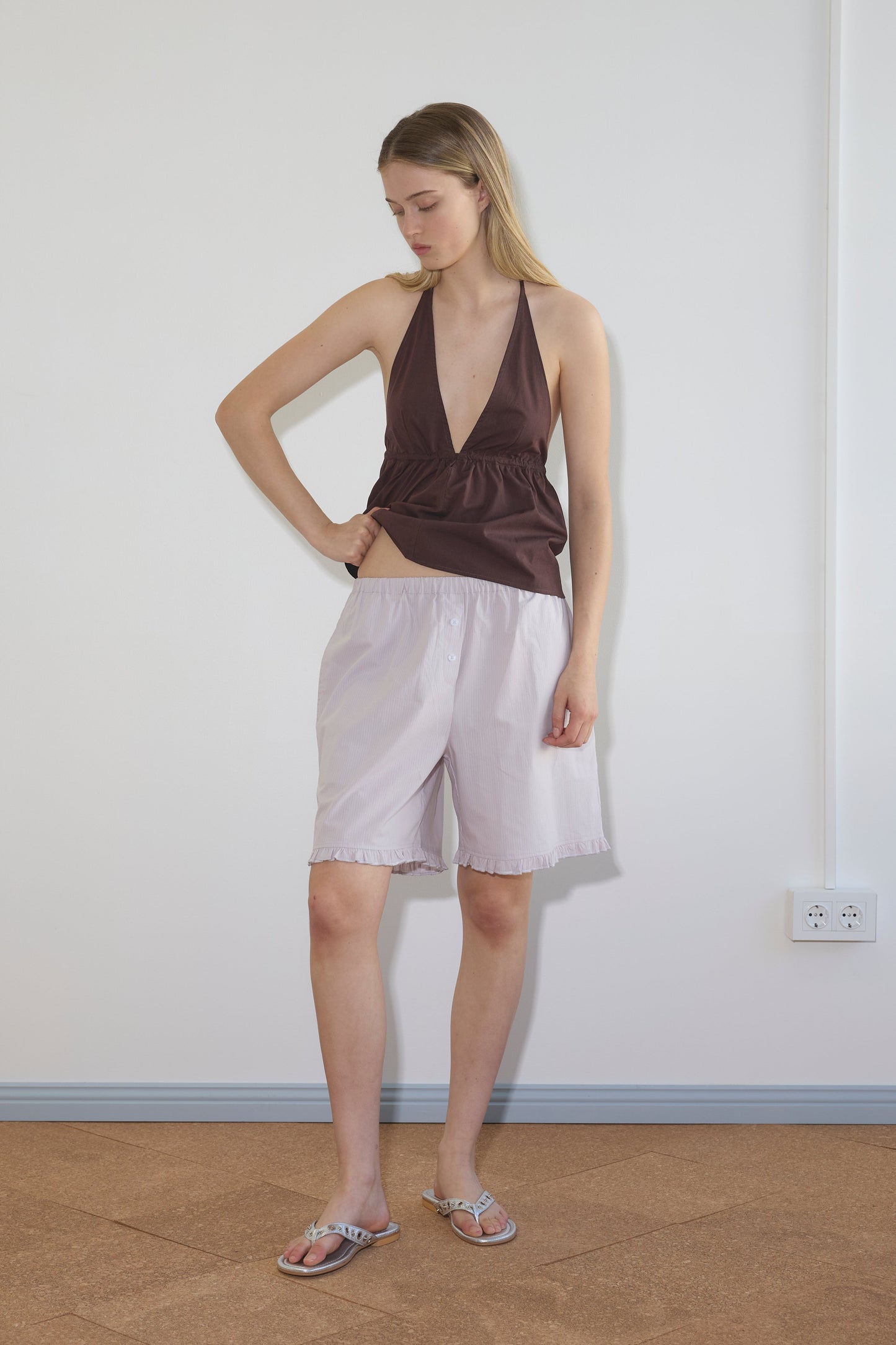 A woman stands indoors in a sleeveless brown halter top and loose, knee-length Flounce Short - Lilac Stripe from Deiji Studios. She looks down with one hand on her hip, paired with flat sandals against a plain background.