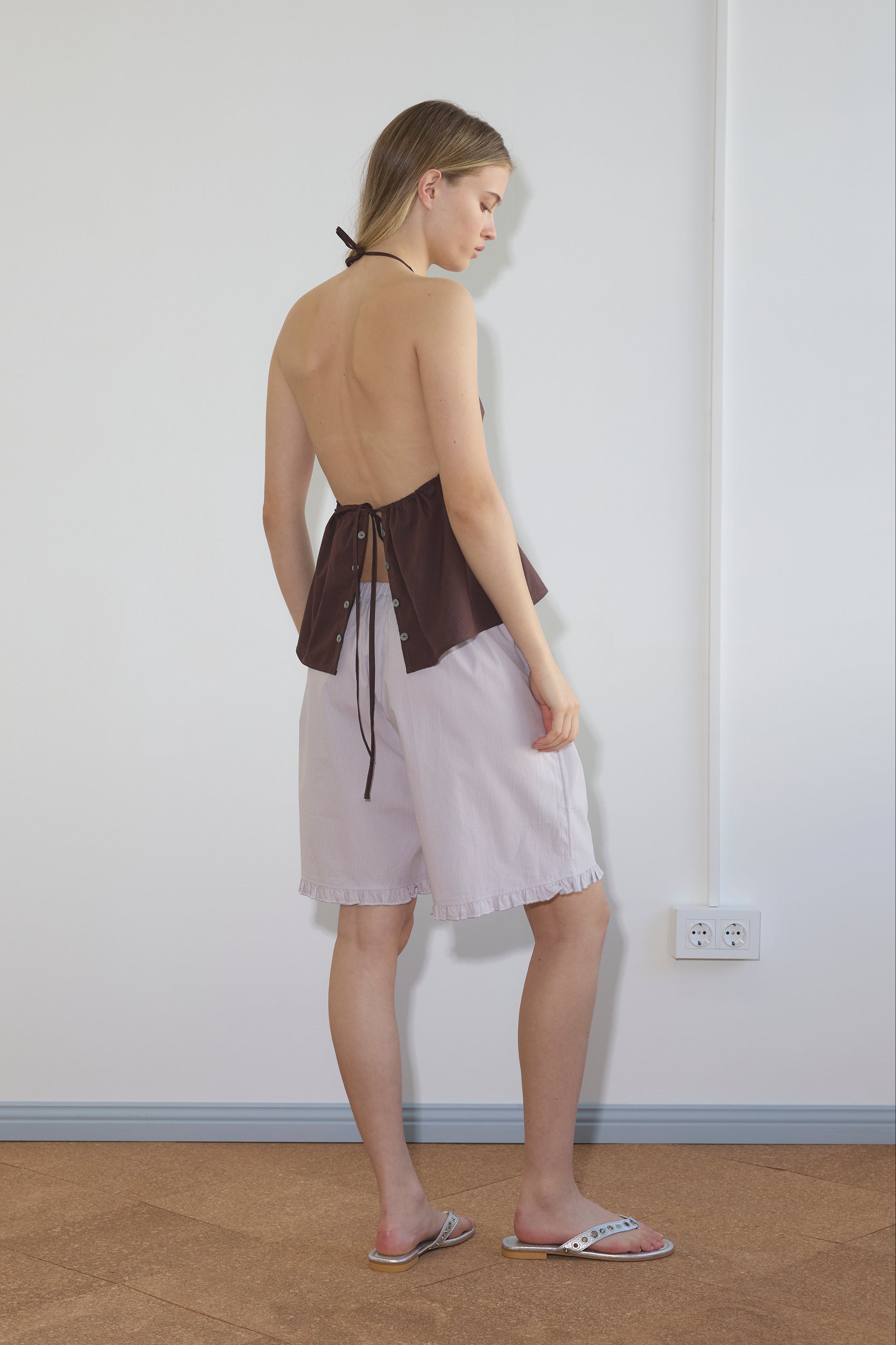 A woman stands indoors, facing sideways, in a brown open-back halter top and Flounce Short - Lilac Stripe by Deiji Studios. She is barefoot except for white flat sandals, with a white electrical outlet visible on the wall behind her.