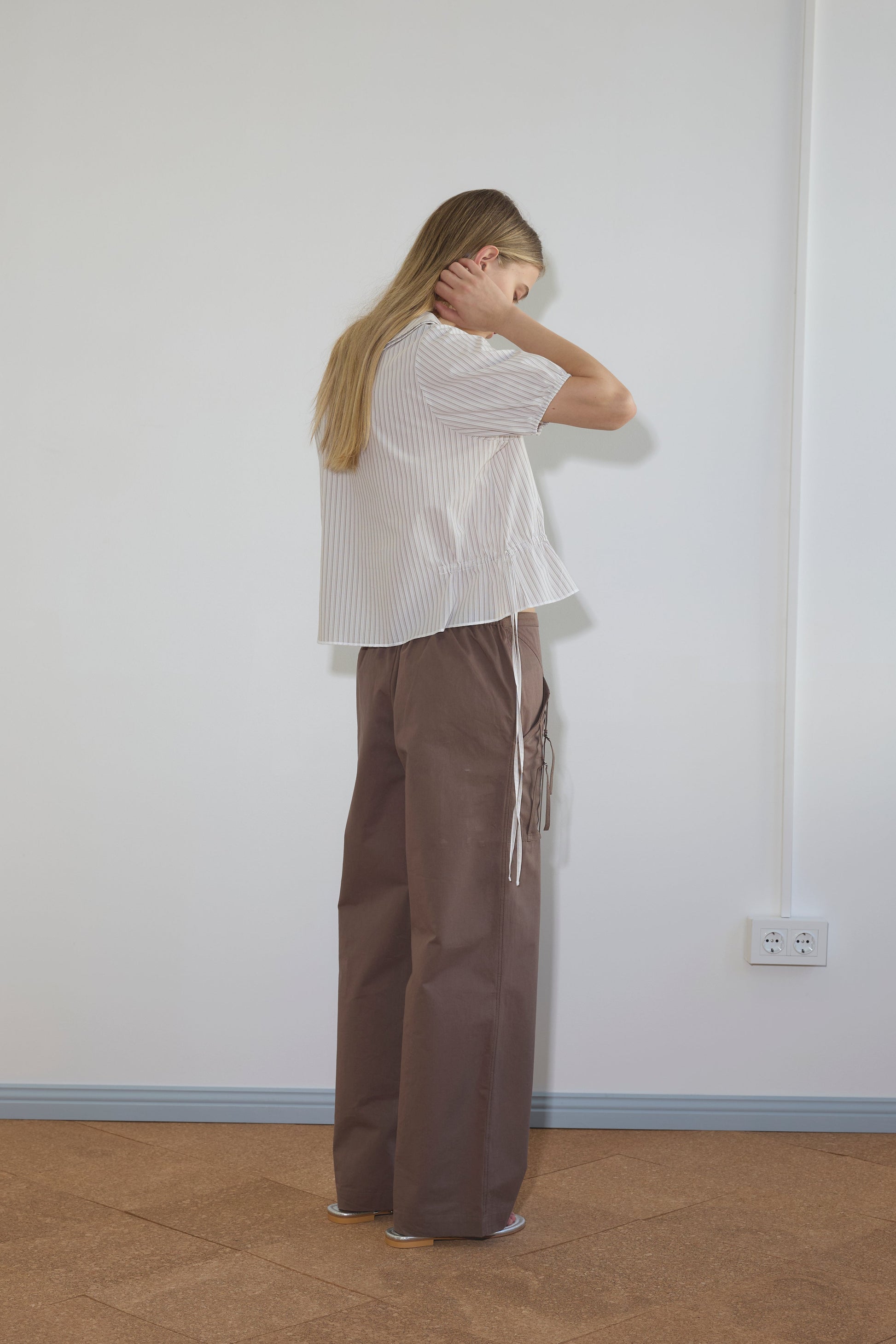 A person with long blonde hair stands facing a wall, wearing the Double Collar Shirt - Gull Stripe in organic cotton. The white, short-sleeve striped blouse is paired with loose brown pants; one hand touches their neck as their face is turned away.