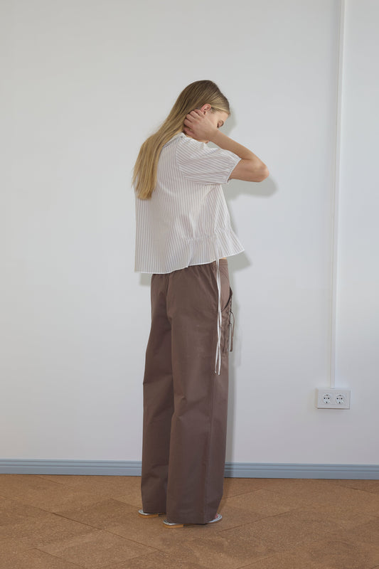 A person with long blonde hair stands facing a wall, wearing the Double Collar Shirt - Gull Stripe in organic cotton. The white, short-sleeve striped blouse is paired with loose brown pants; one hand touches their neck as their face is turned away.