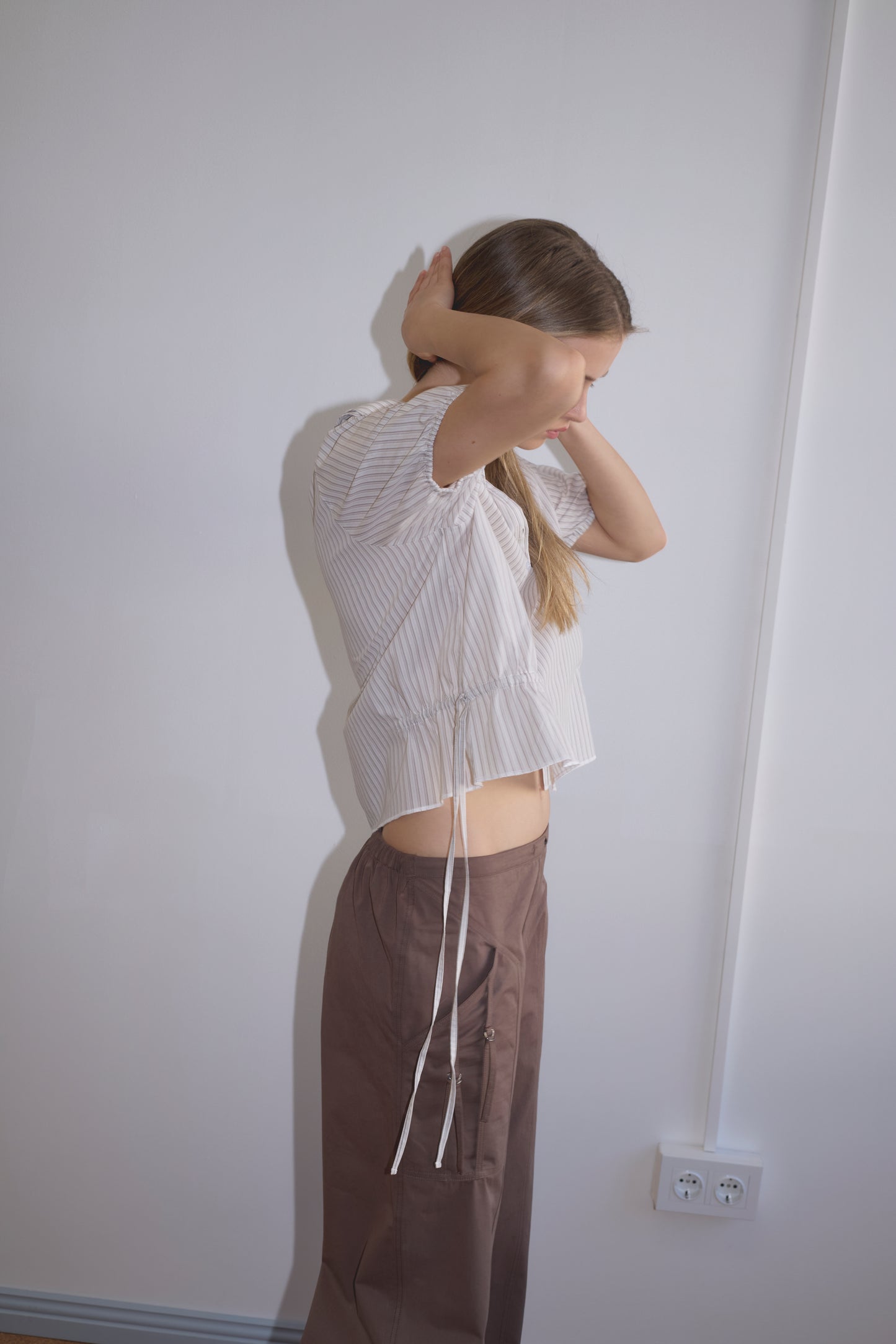 A woman stands sideways against a white wall wearing the Double Collar Shirt - Gull Stripe and brown pants. She raises her arms, partly covering her face with her hands. Two power outlets appear on the wall.