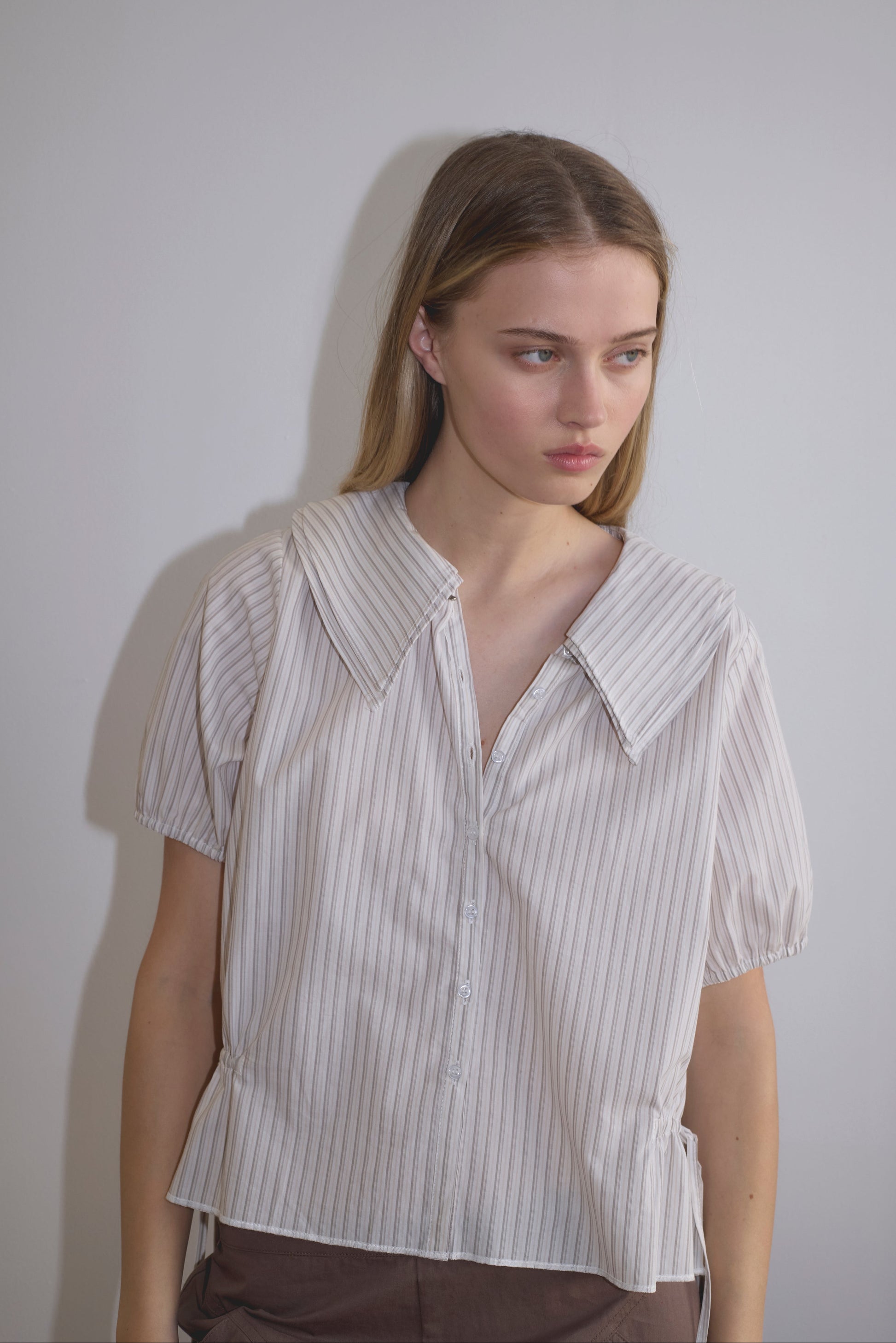 A young woman with long blonde hair stands against a light wall, wearing the Double Collar Shirt - Gull Stripe by Deiji Studios and brown pants. She gazes slightly to the side.