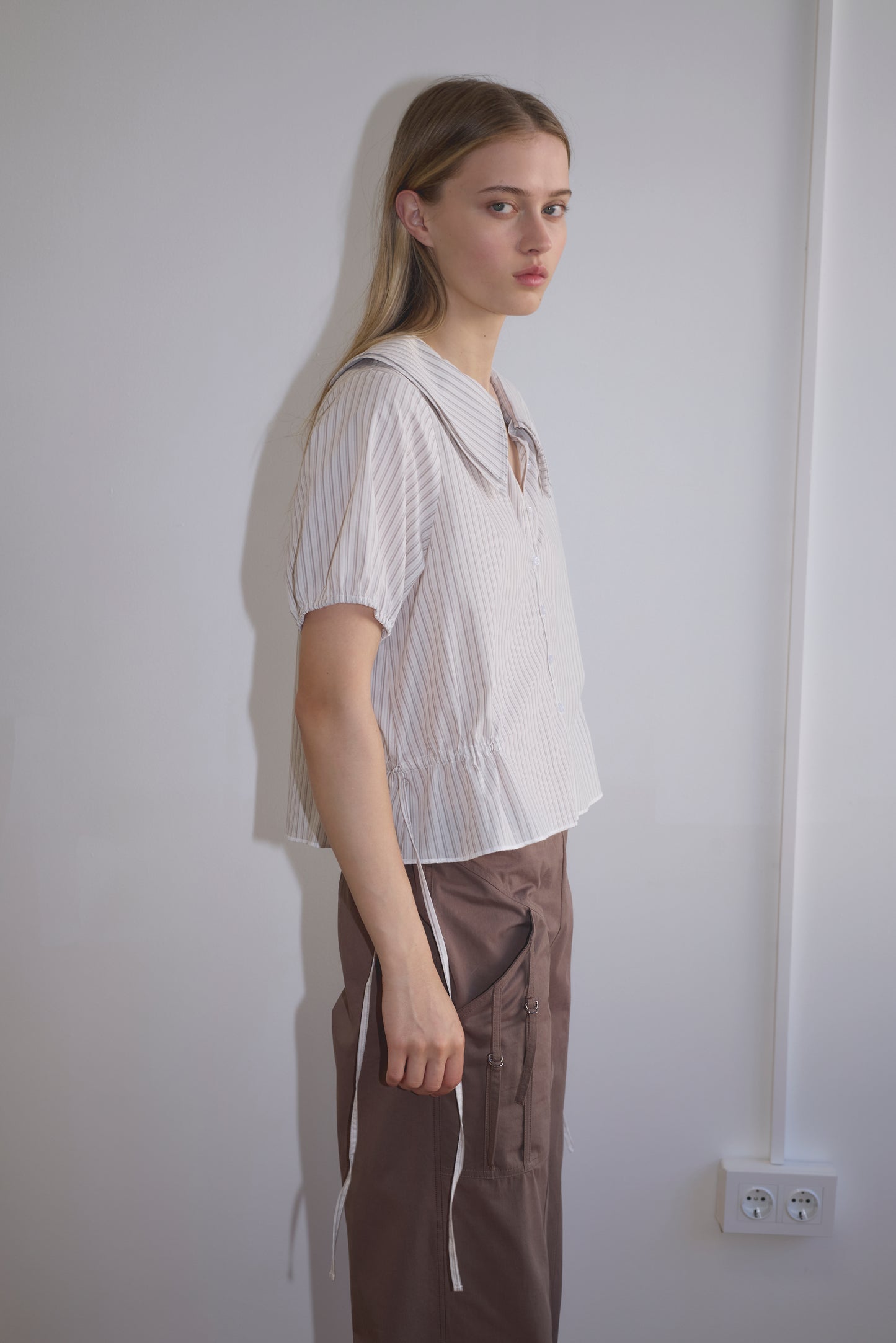 A young woman with long blonde hair stands sideways against a white wall, wearing the Deiji Studios Double Collar Shirt - Gull Stripe and brown pants, looking toward the camera with a neutral expression.