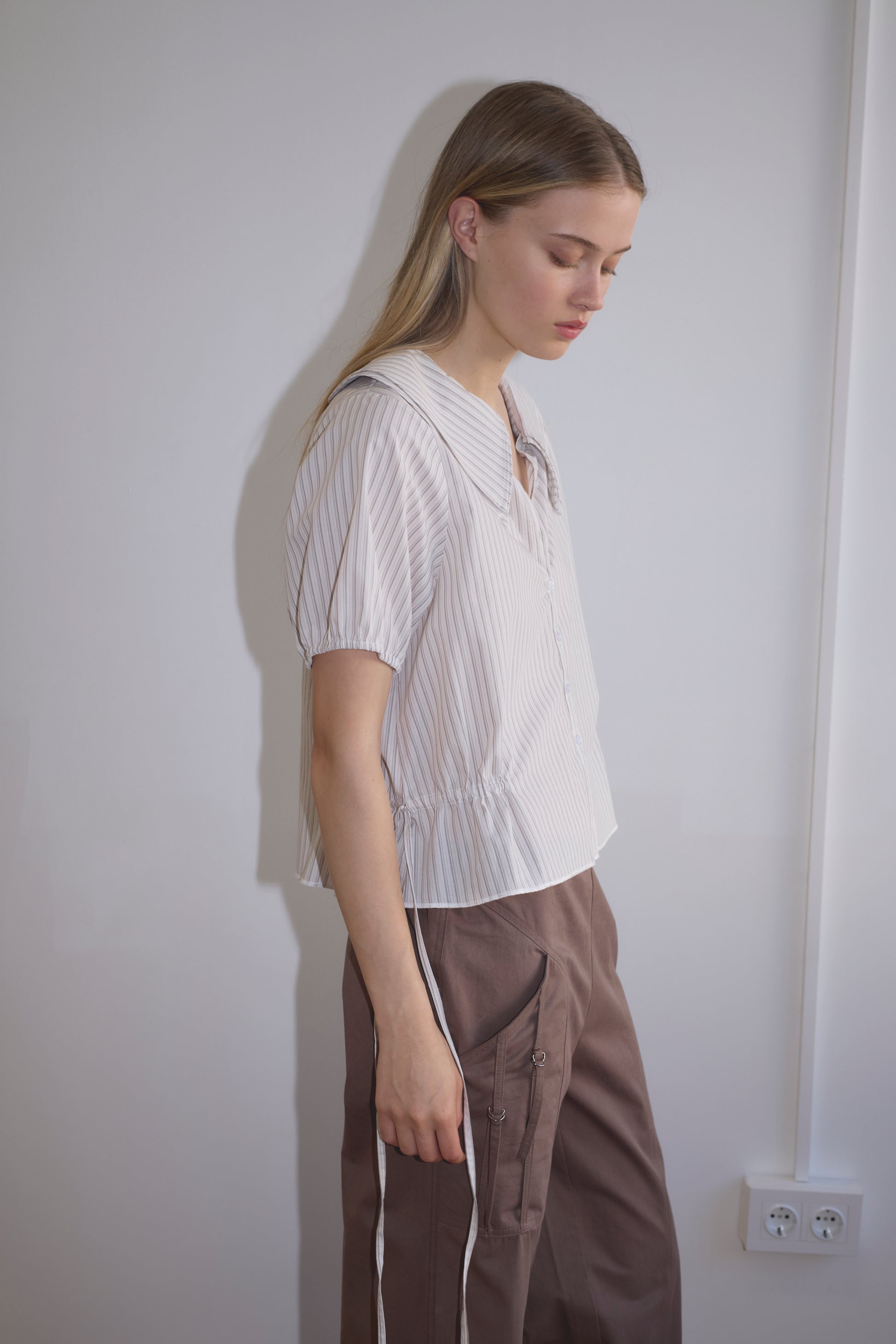 A young woman with long blonde hair stands sideways, looking down. She wears the Double Collar Shirt - Gull Stripe from Deiji Studios and brown pants with white side stripes, against a plain white wall with a visible power outlet.
