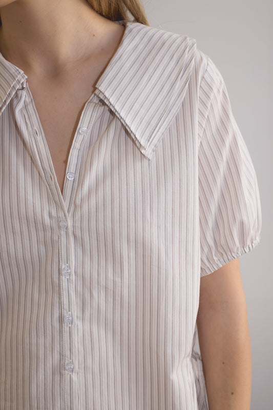 A person wears the Double Collar Shirt - Gull Stripe, a short-sleeved, light-colored, vertically striped organic cotton button-up with a wide collar. Image is cropped to display the upper torso and partial arm.
