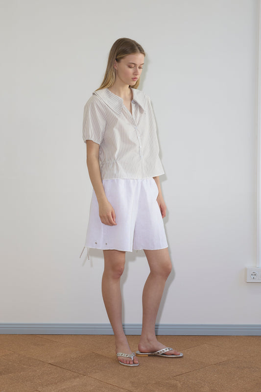 A woman stands indoors by a light wall, wearing the Deiji Studios Double Collar Shirt in Gull Stripe, white knee-length shorts, and open-toe sandals. She has long straight hair and a relaxed expression.