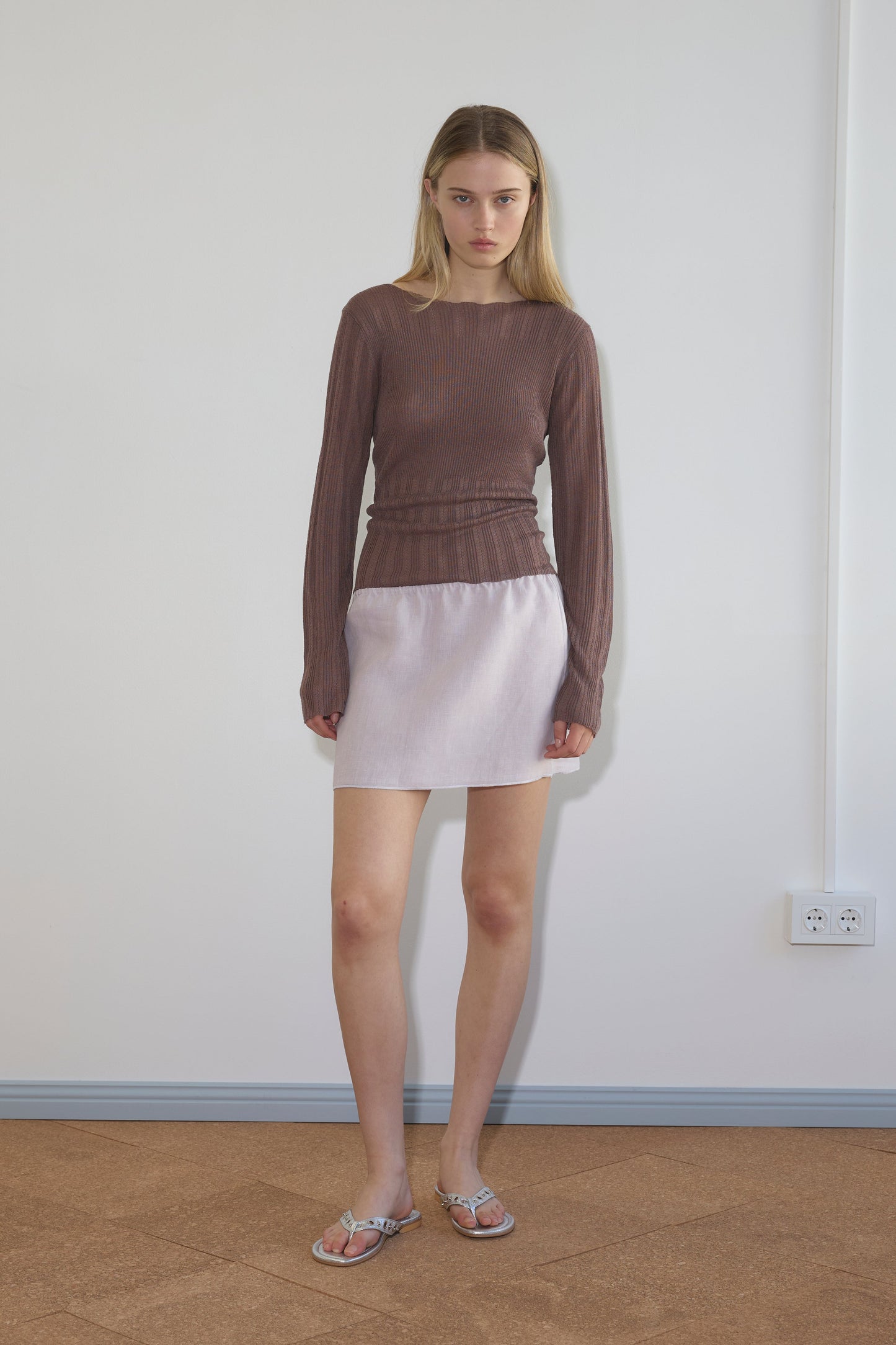 A young woman with straight blonde hair stands indoors against a white wall, wearing a sheer brown long-sleeve top, silver sandals, and the Babylock Skirt in lilac. She has a neutral expression with her arms relaxed at her sides.