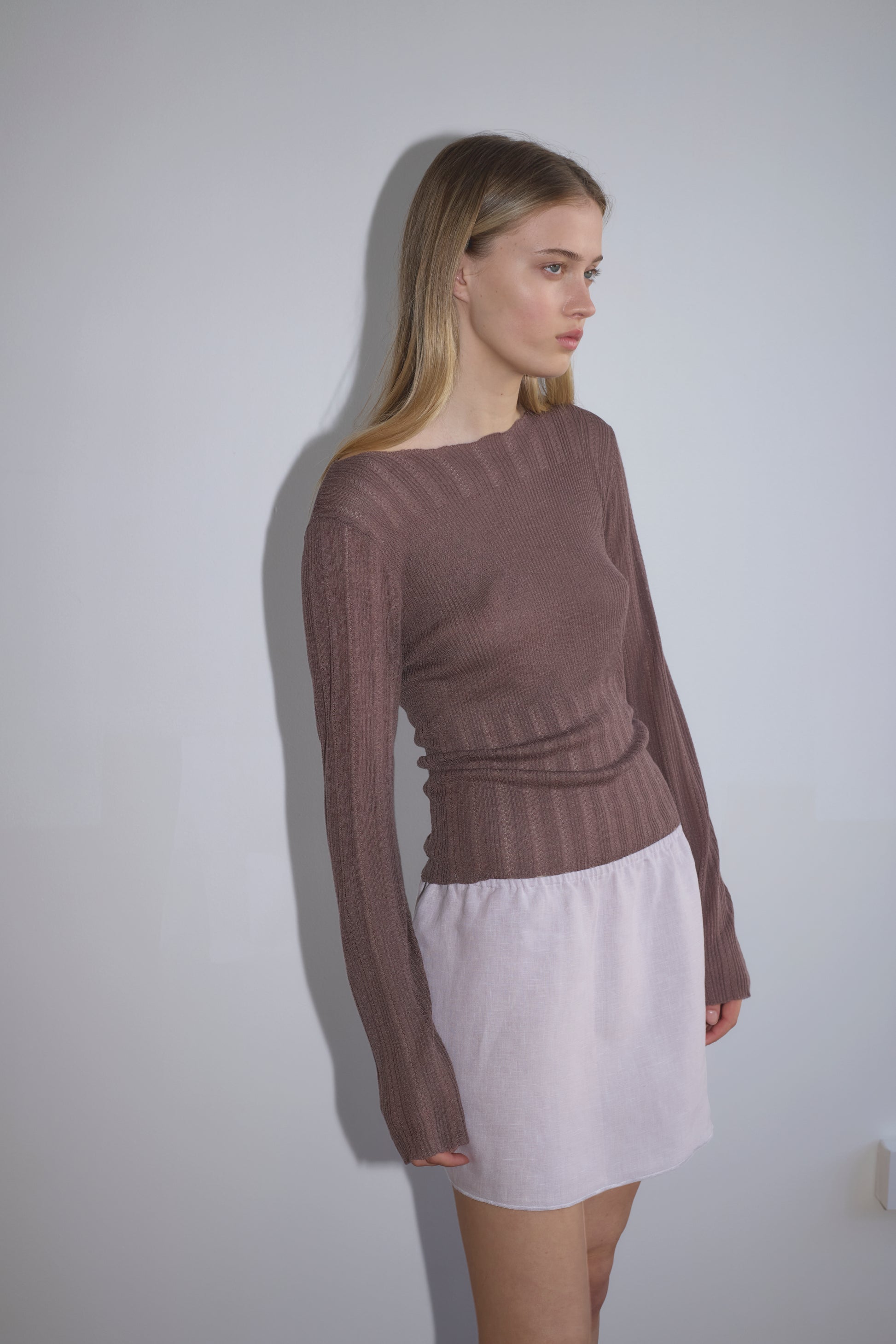 A woman with long blonde hair stands against a light gray wall, wearing a fitted, brown ribbed top and a short Babylock Skirt in lilac. She looks to the side with a neutral expression.