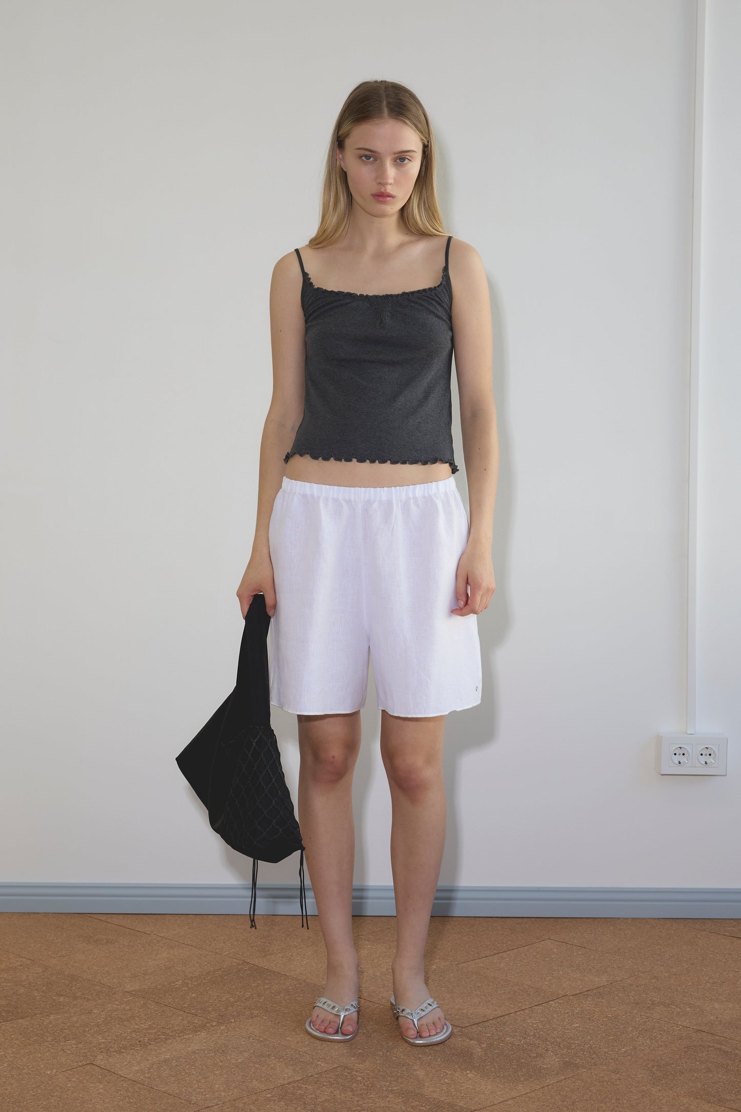 A young woman stands indoors against a plain wall, wearing a dark gray tank top and Deiji Studios Press Stud Shorts in white with silver sandals. She holds a black bag in her left hand, her straight shoulder-length blonde hair framing her face.