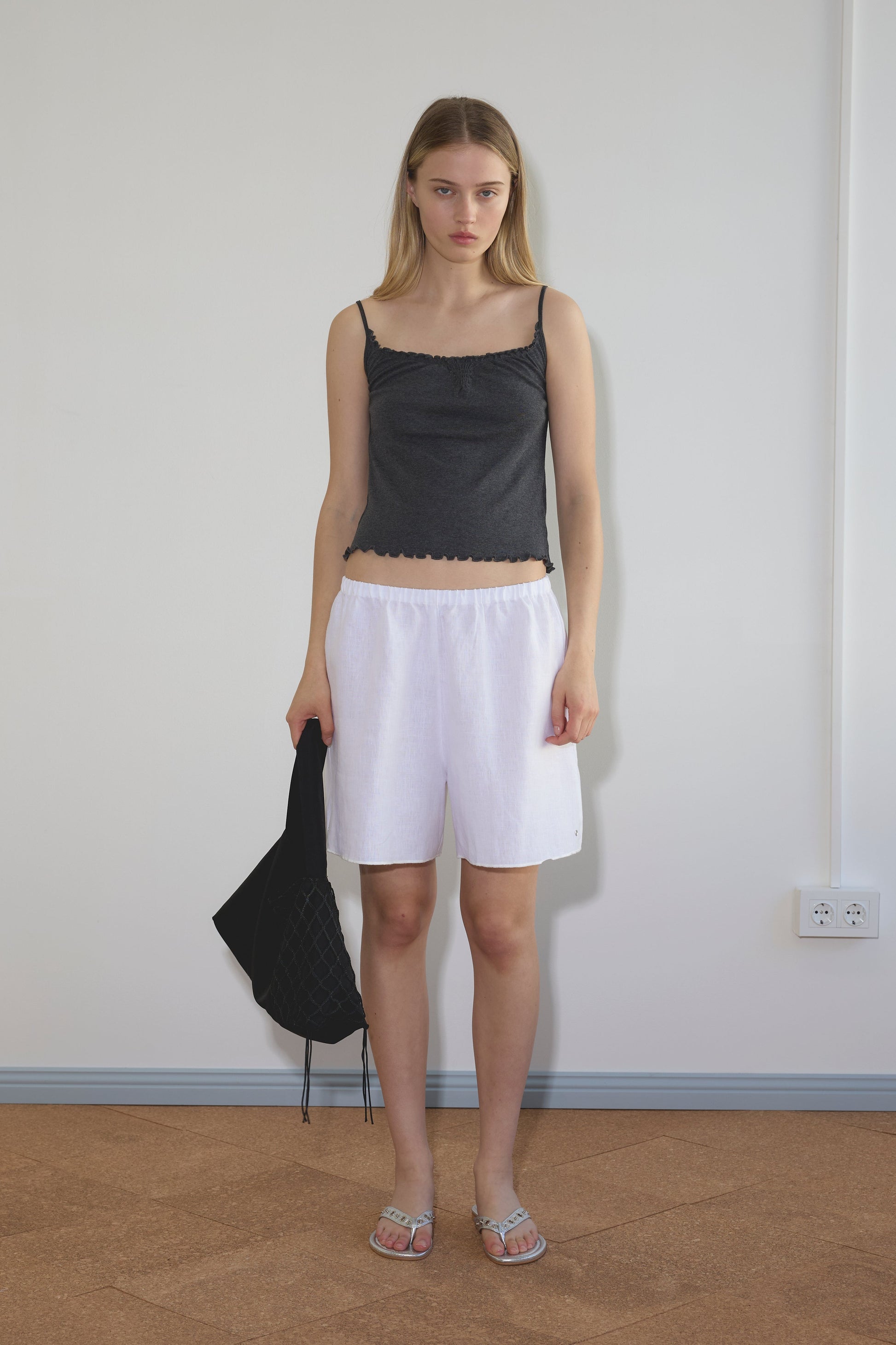 A young woman stands indoors against a plain wall, wearing a dark gray tank top and Deiji Studios Press Stud Shorts in white with silver sandals. She holds a black bag in her left hand, her straight shoulder-length blonde hair framing her face.