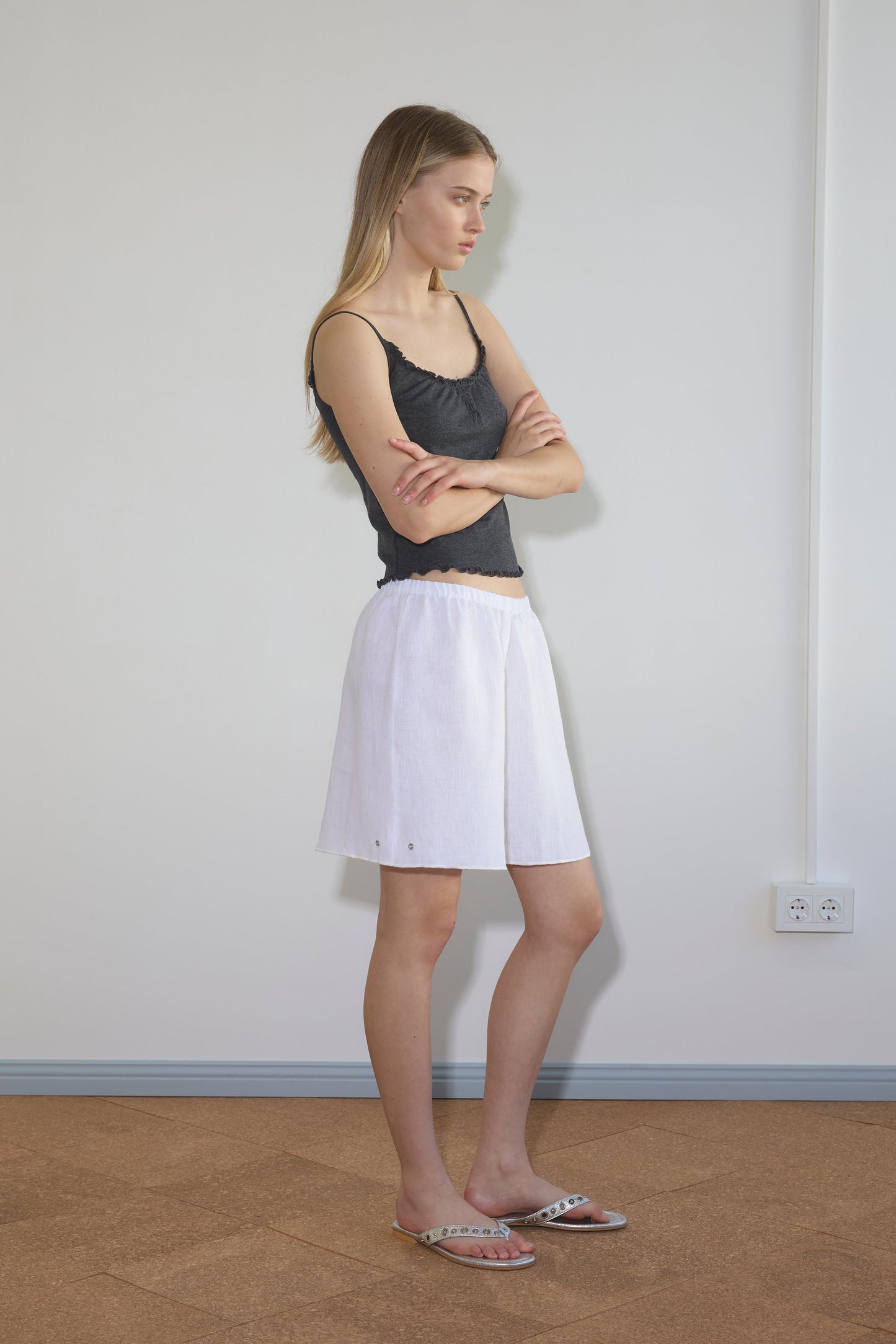 A woman stands sideways indoors against a plain wall, wearing the Press Stud Short in white with a black sleeveless top and sandals. Her long, straight hair is down, and her arms are crossed. A power outlet is visible on the wall behind her.