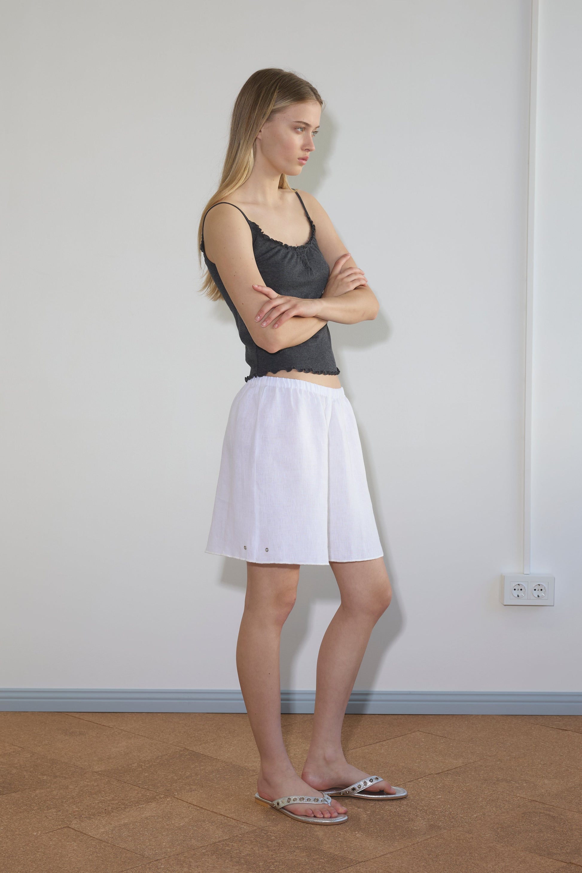 A woman stands sideways indoors against a plain wall, wearing the Press Stud Short in white with a black sleeveless top and sandals. Her long, straight hair is down, and her arms are crossed. A power outlet is visible on the wall behind her.