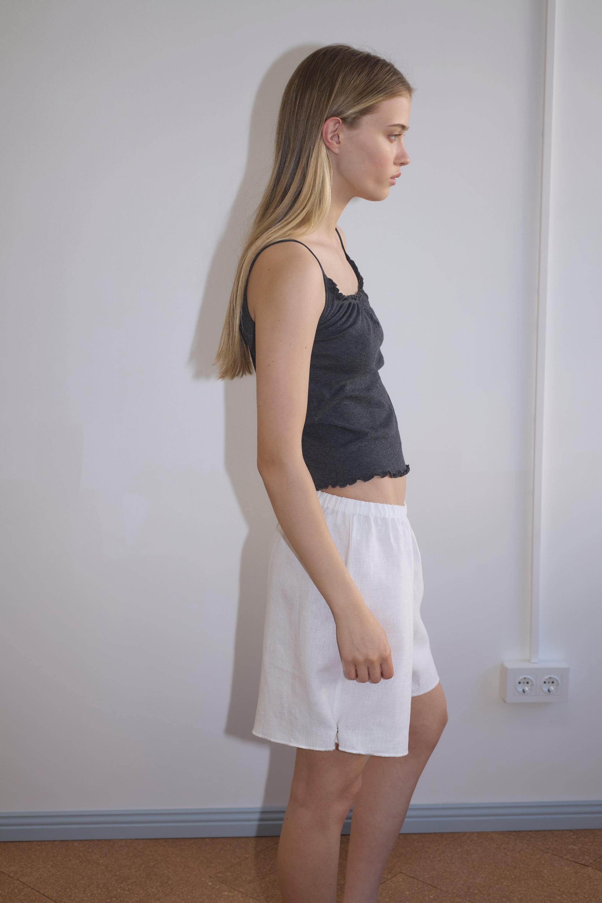 A young woman with long blonde hair stands in profile against a plain wall, wearing a black sleeveless top and the white Press Stud Short by Deiji Studios. A power outlet is visible on the wall behind her.