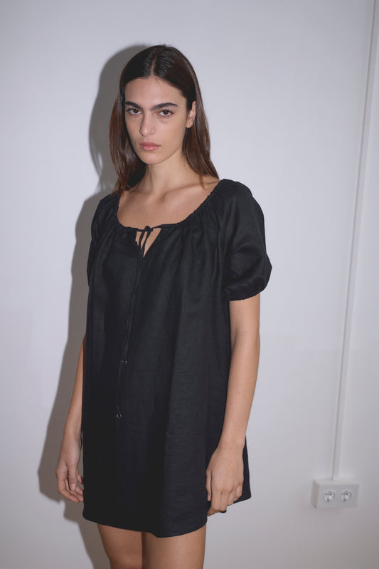 A person with long brown hair stands against a white wall, wearing the Smock Dress - Black, a loose-fitting French linen dress with short sleeves and a tie neckline, evoking Deiji Studios’ iconic style.