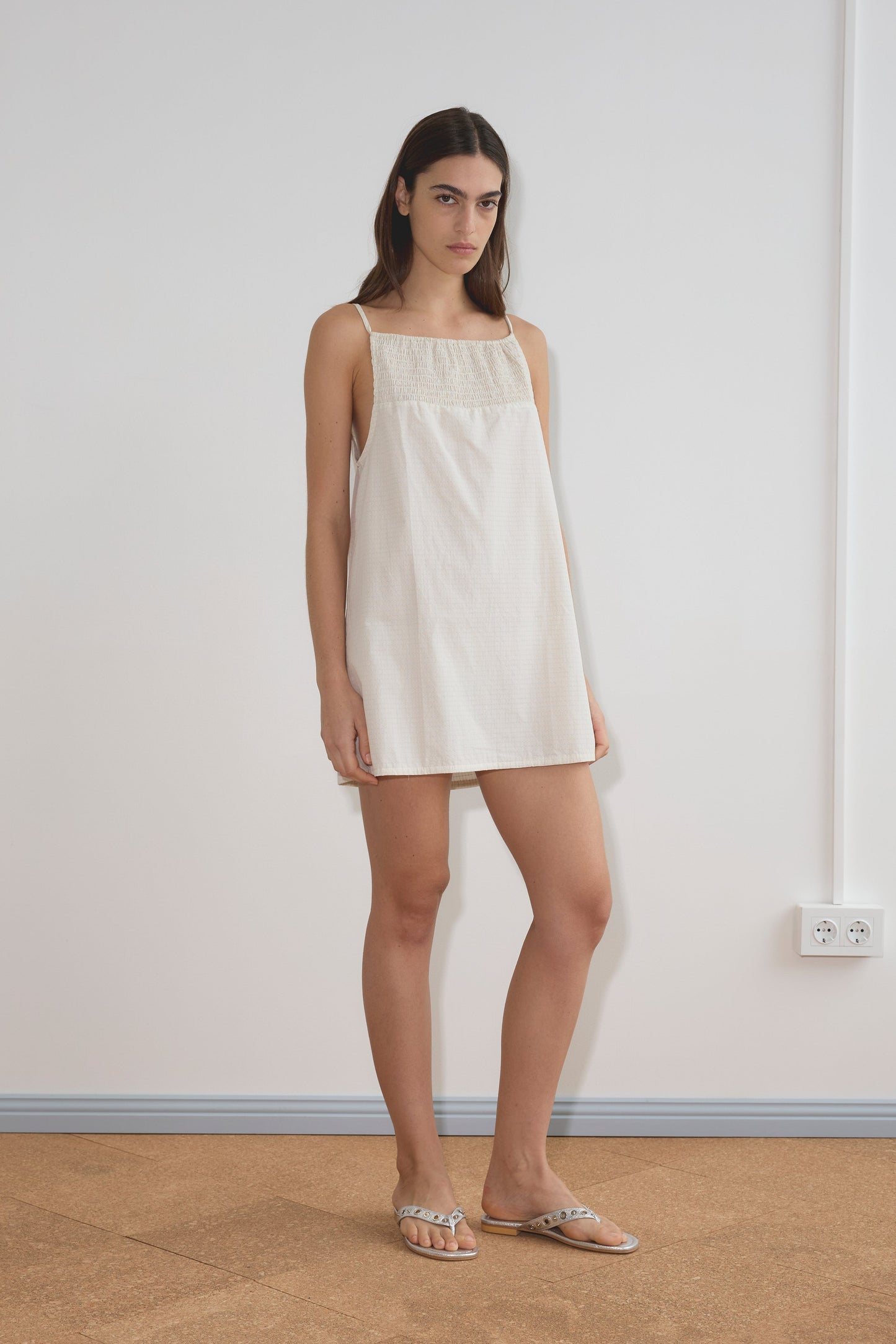 A woman stands indoors against a plain white wall, wearing the Shirring Mini Dress - Bone Check by Deiji Studios and flat sandals. She has straight dark hair and a neutral expression.