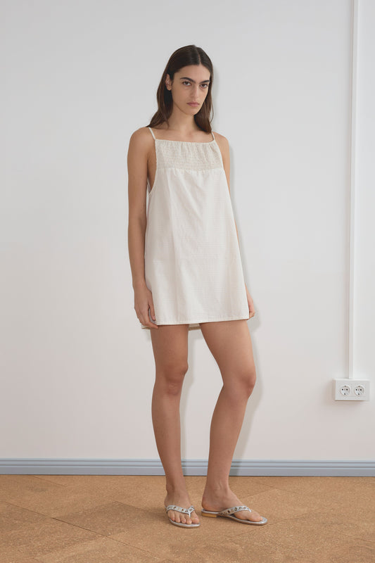 A woman stands indoors against a plain white wall, wearing the Shirring Mini Dress - Bone Check by Deiji Studios and flat sandals. She has straight dark hair and a neutral expression.