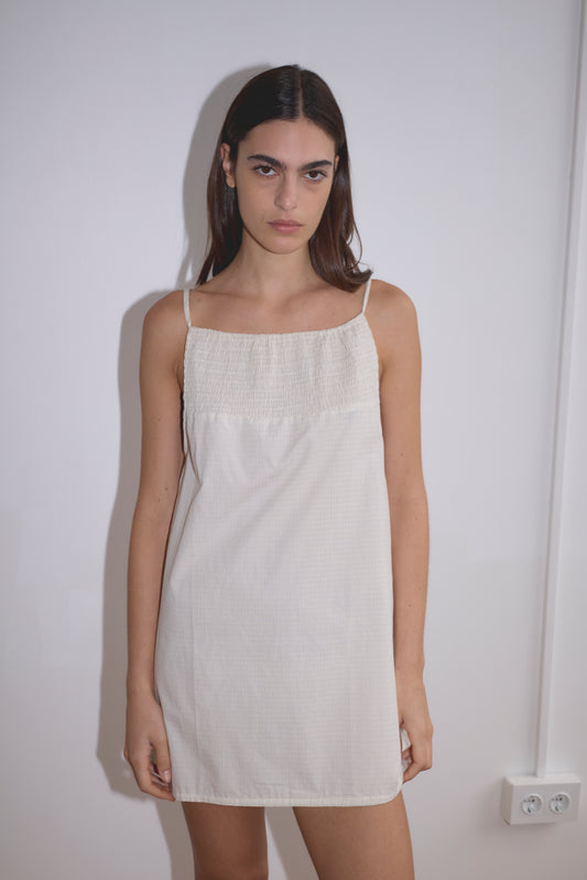 A woman with long dark hair stands against a white wall, wearing the Shirring Mini Dress - Bone Check by Deiji Studios. The dress is simple, sleeveless with thin straps. Her expression is neutral and her arms rest by her sides.