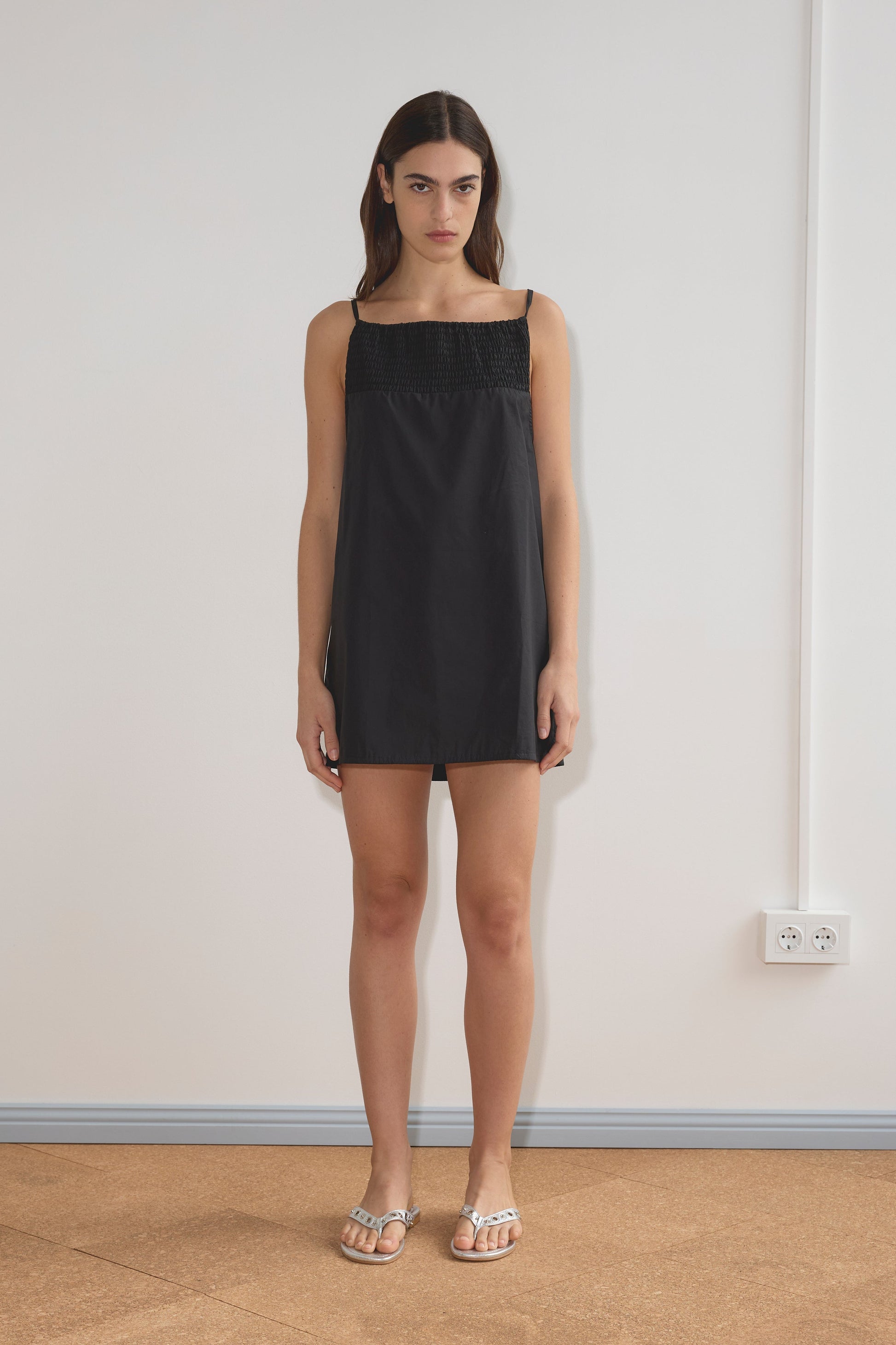 A woman with straight, dark hair and a neutral expression stands indoors against a plain white wall, wearing the Shirring Mini Dress - Black by Deiji Studios with a textured upper panel and metallic sandals.