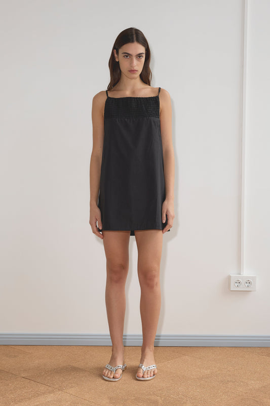 A woman with straight, dark hair and a neutral expression stands indoors against a plain white wall, wearing the Shirring Mini Dress - Black by Deiji Studios with a textured upper panel and metallic sandals.
