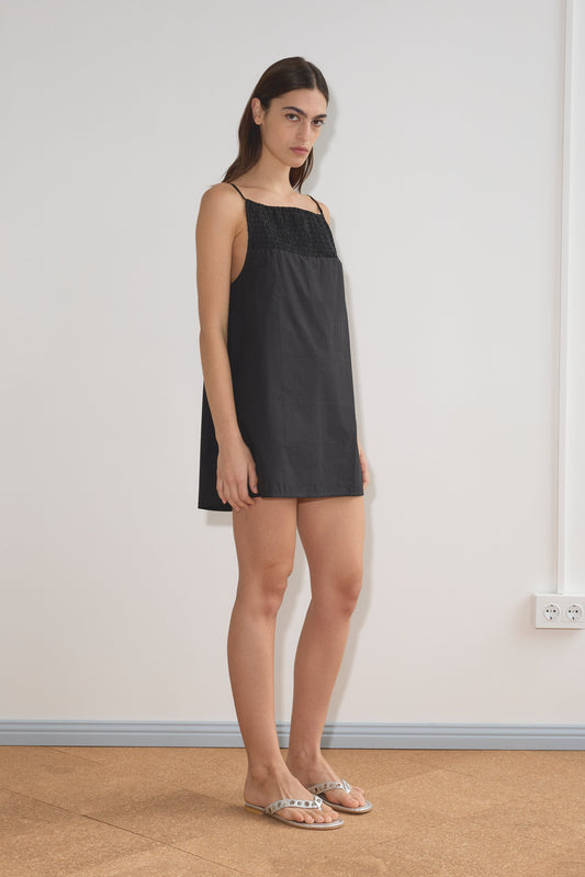 A woman with straight brown hair wears the Shirring Mini Dress - Black with flat sandals, standing indoors on a tan floor against a plain white wall.