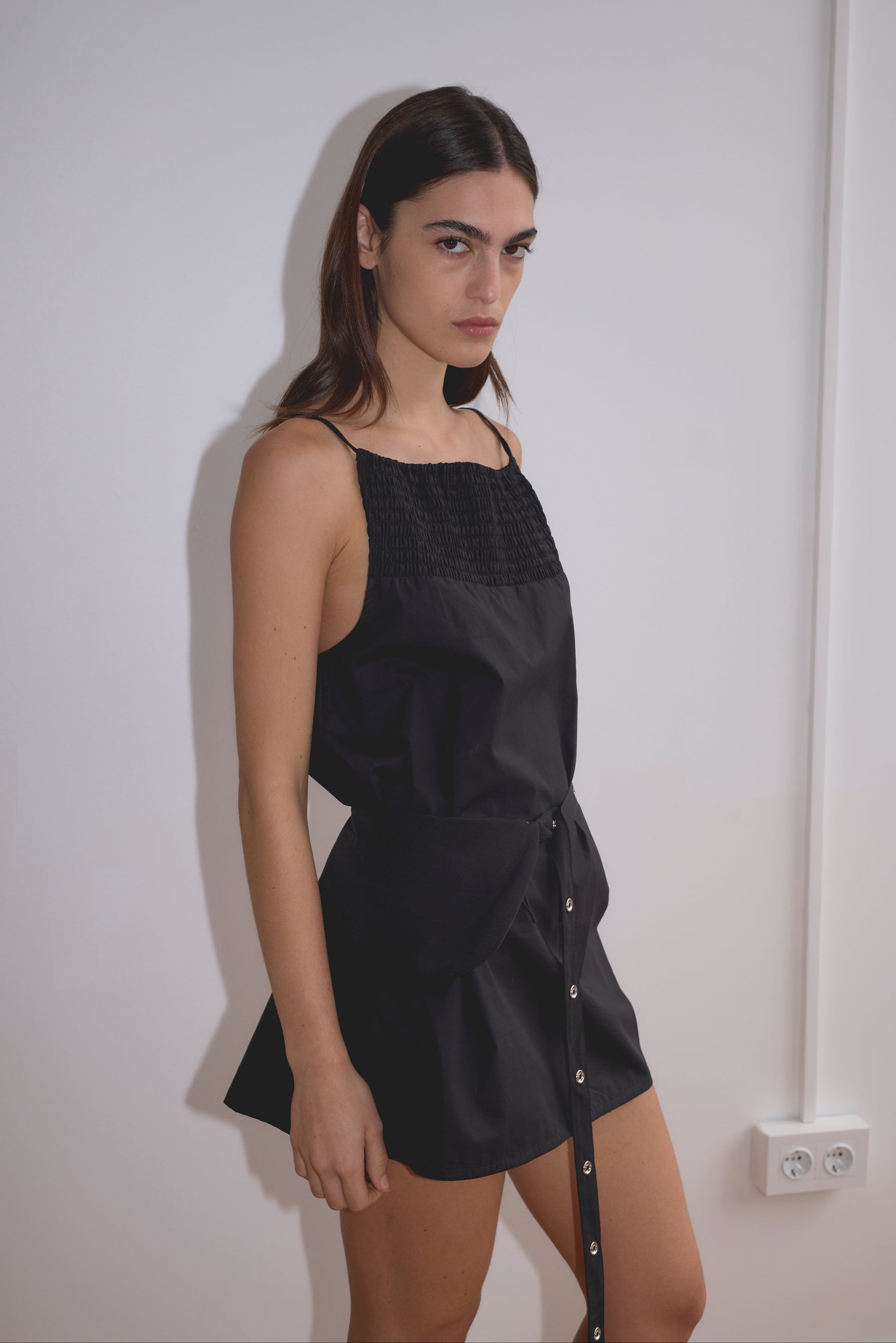 A woman with long dark hair stands against a white wall wearing the Shirring Mini Dress - Black by Deiji Studios, featuring a belted waist and button details. She gazes at the camera with a neutral expression; an electrical outlet is visible nearby.