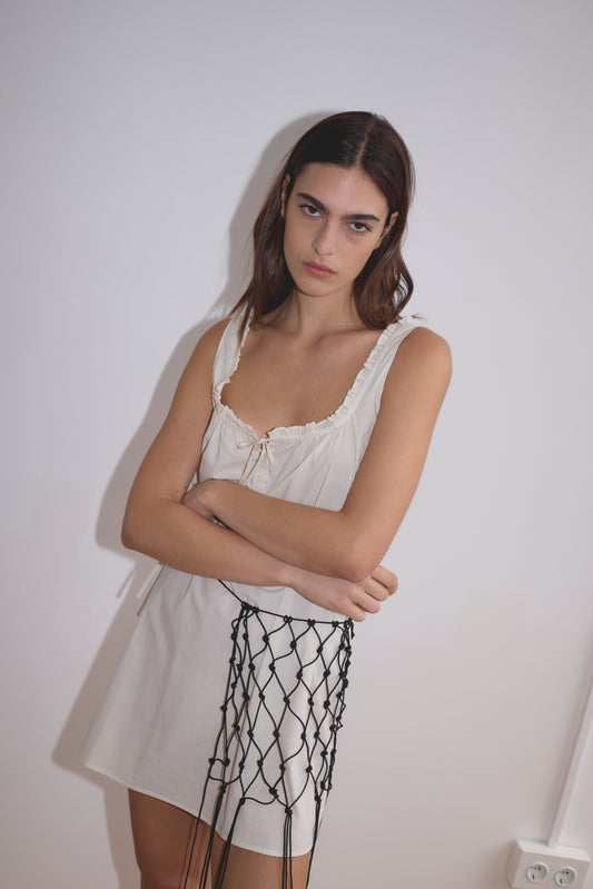 A woman with long brown hair stands against a white wall, wearing the Button Frill Mini Dress in Stone Stripe by Deiji Studios. The organic cotton dress features ruffled edges. She stands with arms crossed, looking serious, with a black netted accessory at her hip.