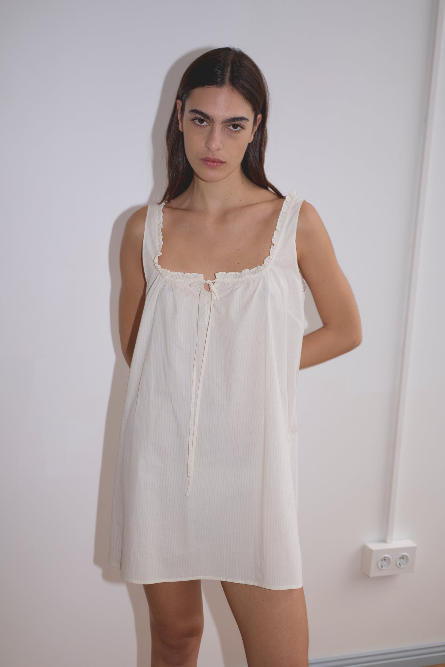 A woman with long dark hair stands against a white wall, wearing the Button Frill Mini Dress in Stone Stripe by Deiji Studios. The sleeveless, loose-fitting organic cotton dress features a tie at the neckline. She looks directly at the camera, expression neutral.
