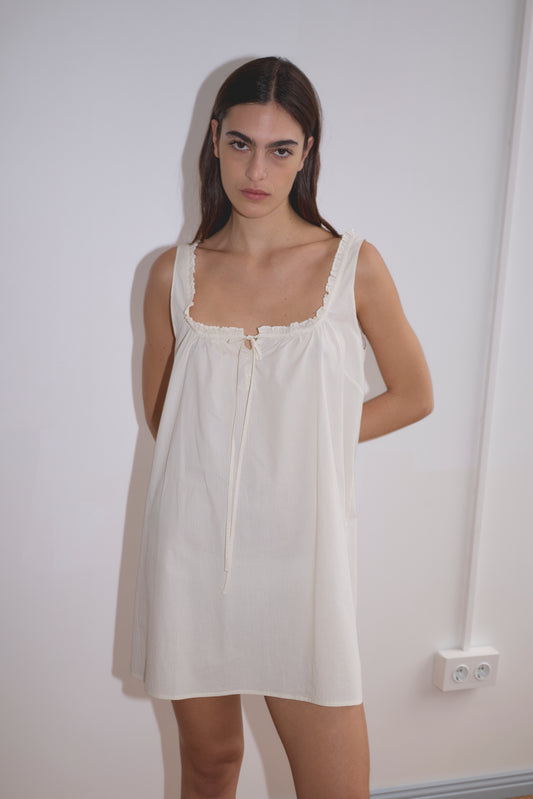 A woman with long dark hair stands against a white wall, wearing the Button Frill Mini Dress in Stone Stripe by Deiji Studios. The sleeveless, loose-fitting organic cotton dress features a tie at the neckline. She looks directly at the camera, expression neutral.