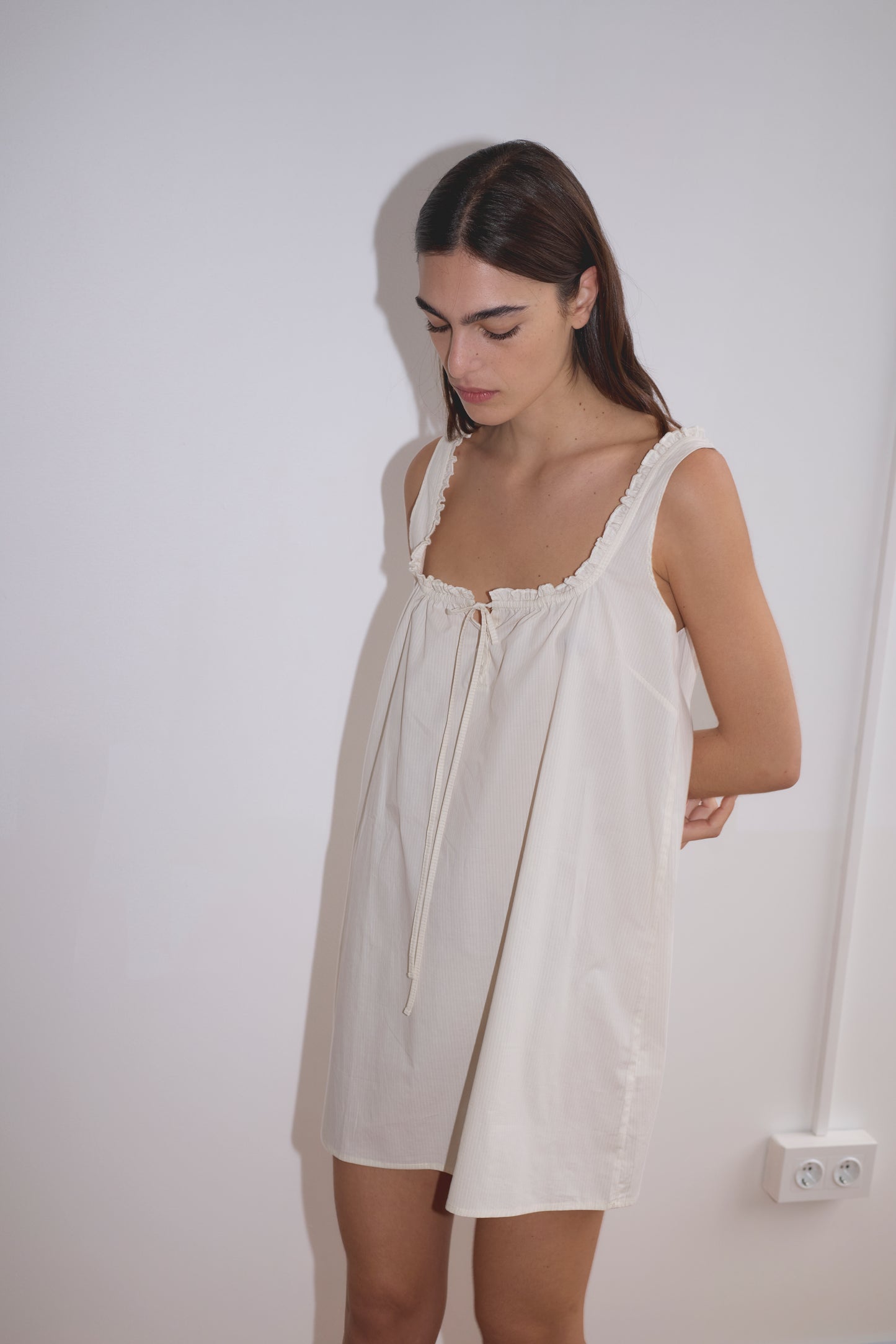 A woman with long dark hair stands against a white wall, wearing the Button Frill Mini Dress - Stone Stripe. Featuring a ruffled neckline and front tie, she looks down with a calm expression in this effortlessly chic look.