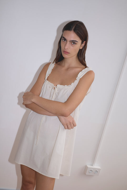 A woman with long brown hair, wearing the Button Frill Mini Dress in Stone Stripe, stands against a plain white wall with her arms crossed and a neutral expression. An electrical socket is visible on the wall behind her.