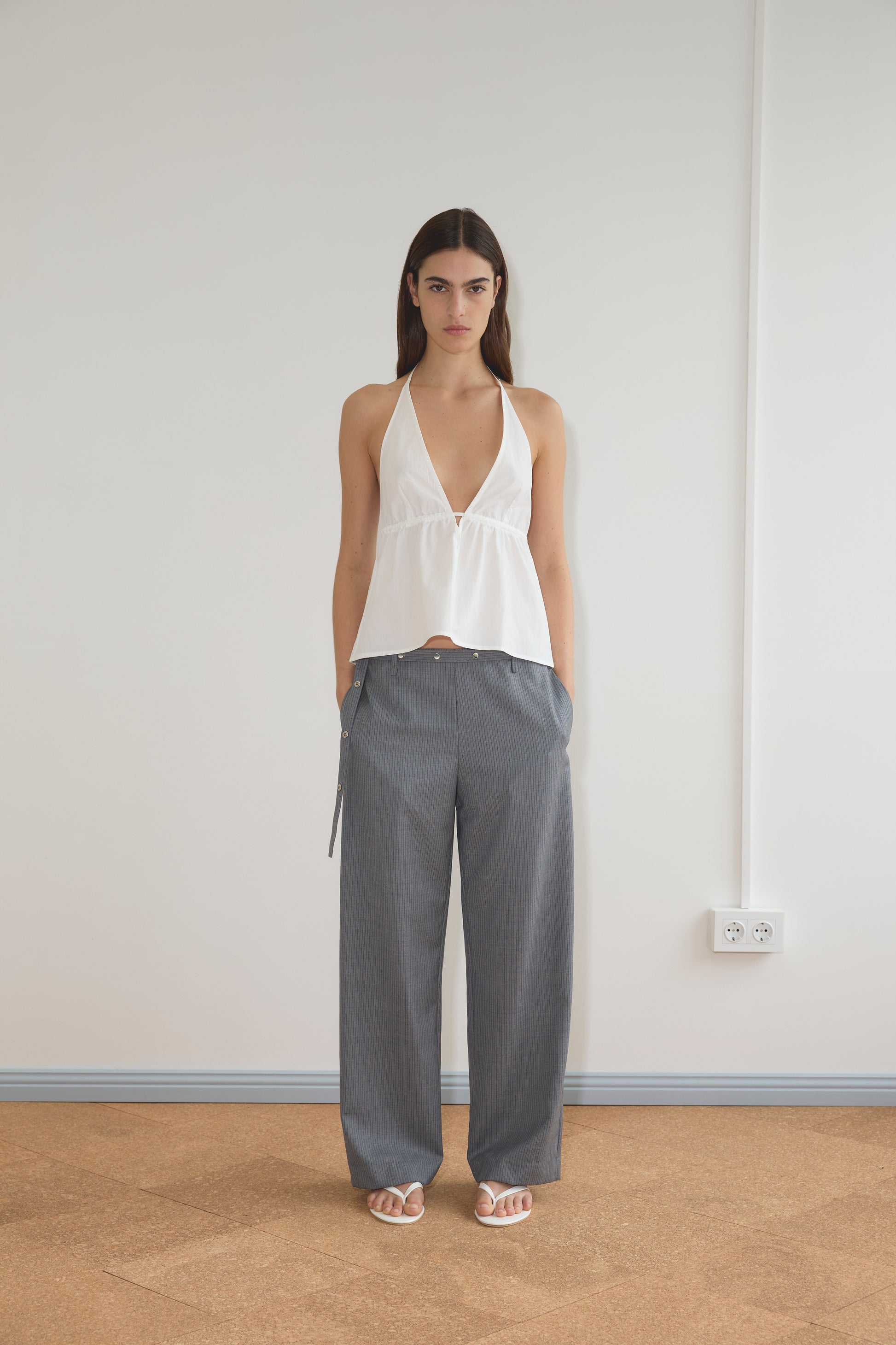 A woman stands indoors against a plain wall, wearing the Button Halter - White and loose-fitting gray pinstripe trousers from Deiji Studios, paired with white sandals. Her hands are in her pockets as she looks at the camera.
