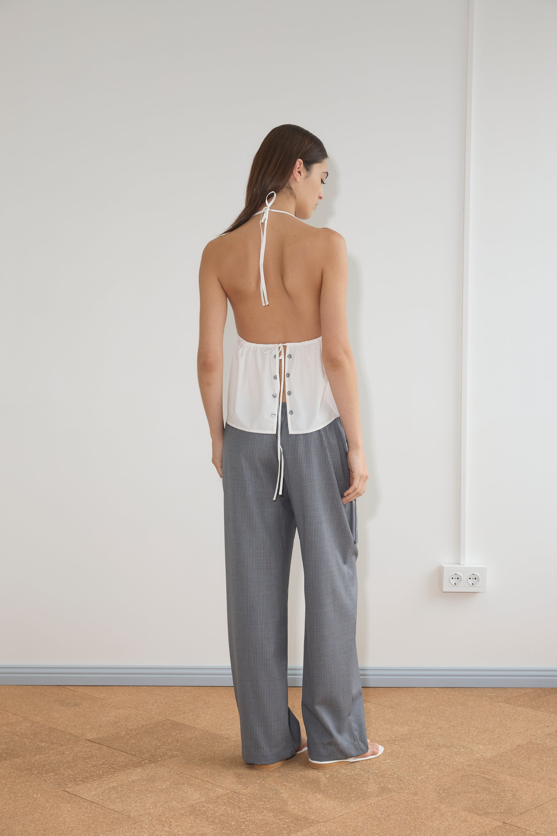 A woman with long brown hair stands indoors on a light brown floor, facing a white wall. She wears the Deiji Studios Button Halter - White, an open-back halter top, styled with loose gray organic cotton pants.