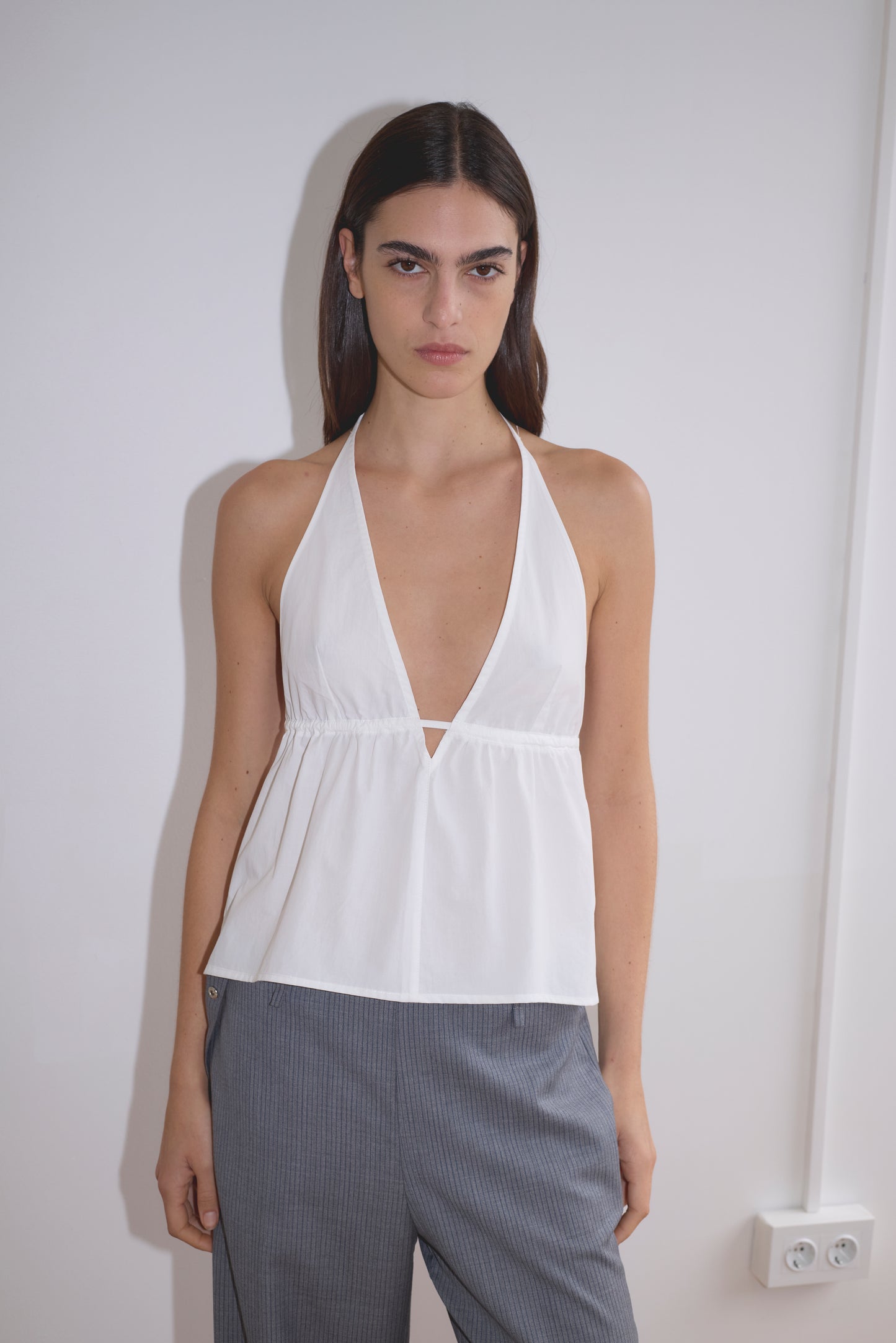 A woman with long dark hair wears the white Button Halter from Deiji Studios, featuring a sleeveless design and deep V neckline, paired with loose gray pants, as she stands against a plain white wall.
